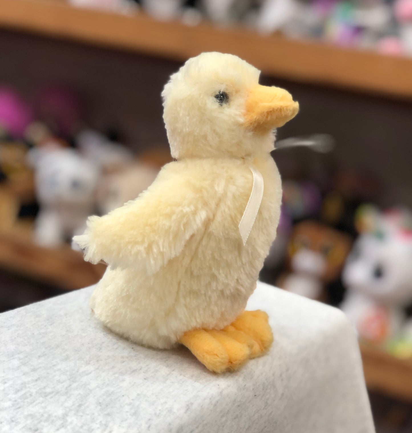 Soft yellow baby duck plush toy with orange feet and bill, wearing a ribbon, displayed on white surface in store