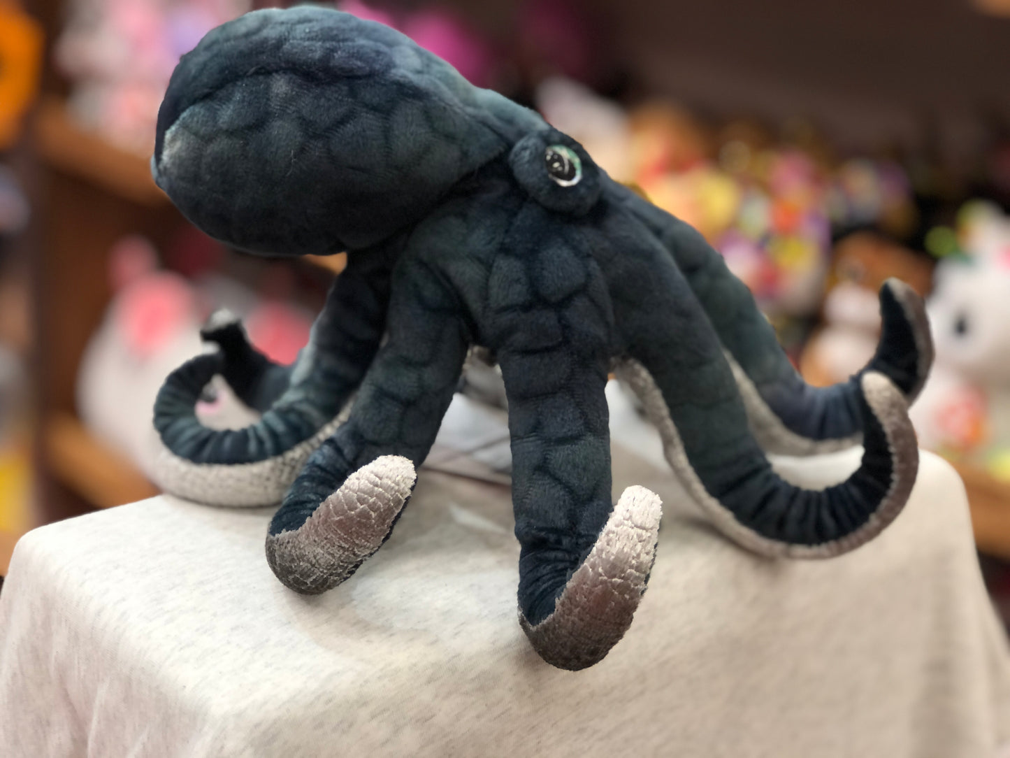 Realistic dark blue octopus plush toy with textured body and curled tentacles, displayed on white surface in toy store
