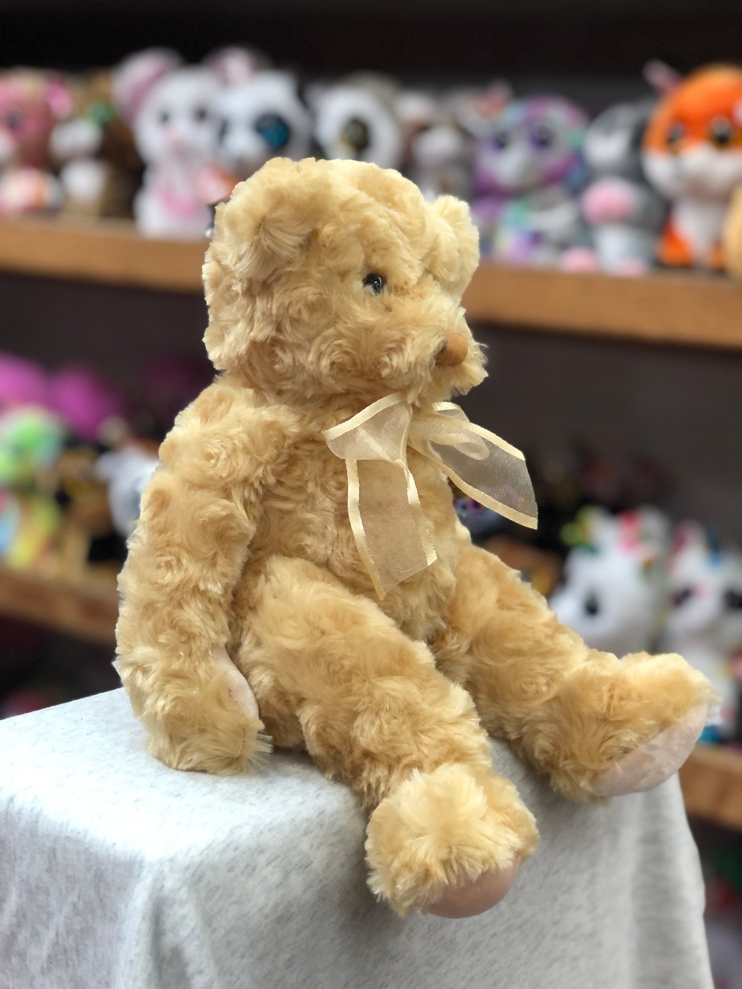 Soft golden Douglas Tender Teddy Bear plush toy with ribbon bow, seated on display with other stuffed animals in background