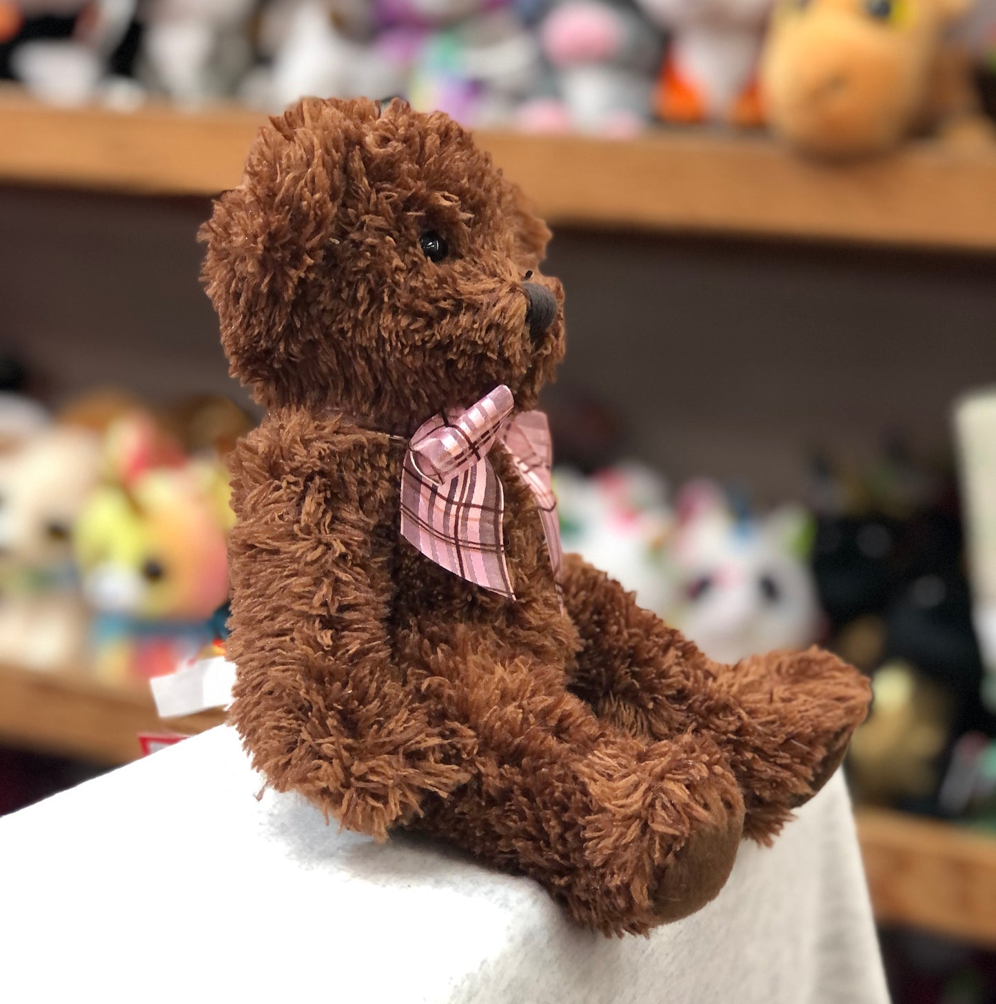 Soft chocolate brown teddy bear plush with pink ribbon bow, sitting on display among other stuffed toys