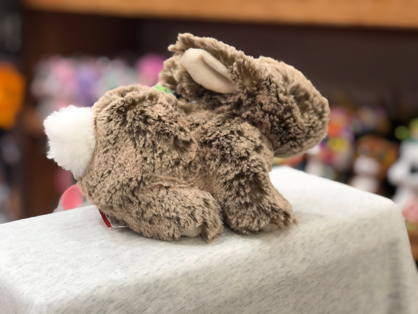 Soft mocha bunny plush toy with long ears and fluffy white tail, displayed on white surface with colorful toys in background