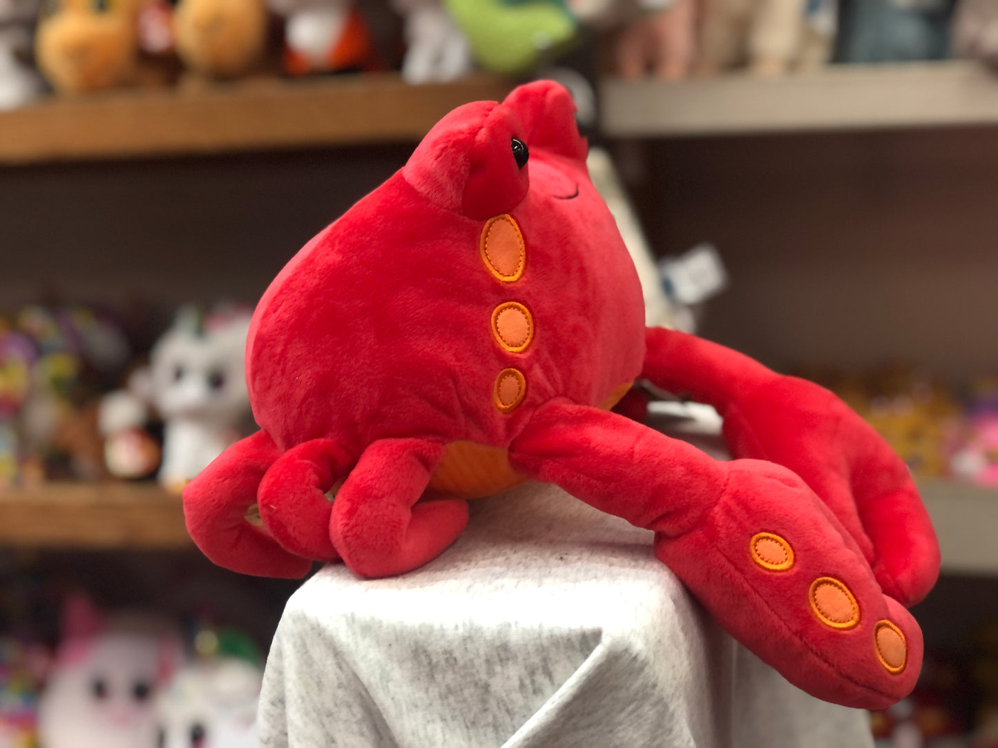 Gund Sydney Crab plush toy, bright red with orange spots, smiling, soft and huggable, displayed on shelf with other stuffed animals