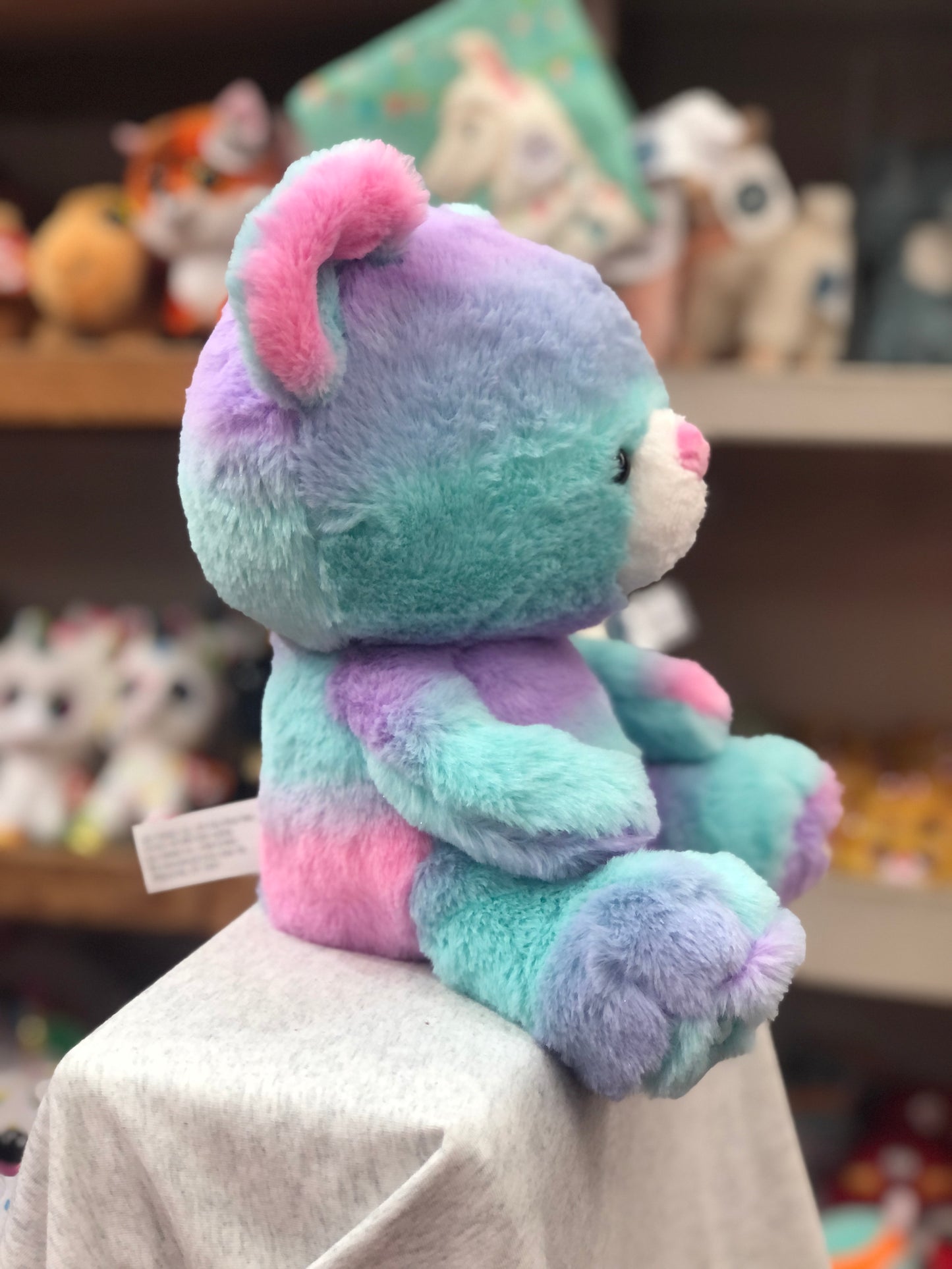 Colorful Gund Kai tie-dye teddy bear plush toy with oversized head, sitting on display in store