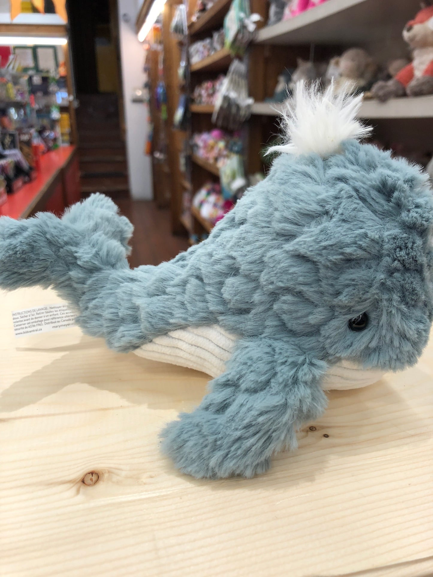 Soft blue Mary Meyer Puttling Whale plush toy with white tuft on display in toy store, surrounded by other stuffed animals