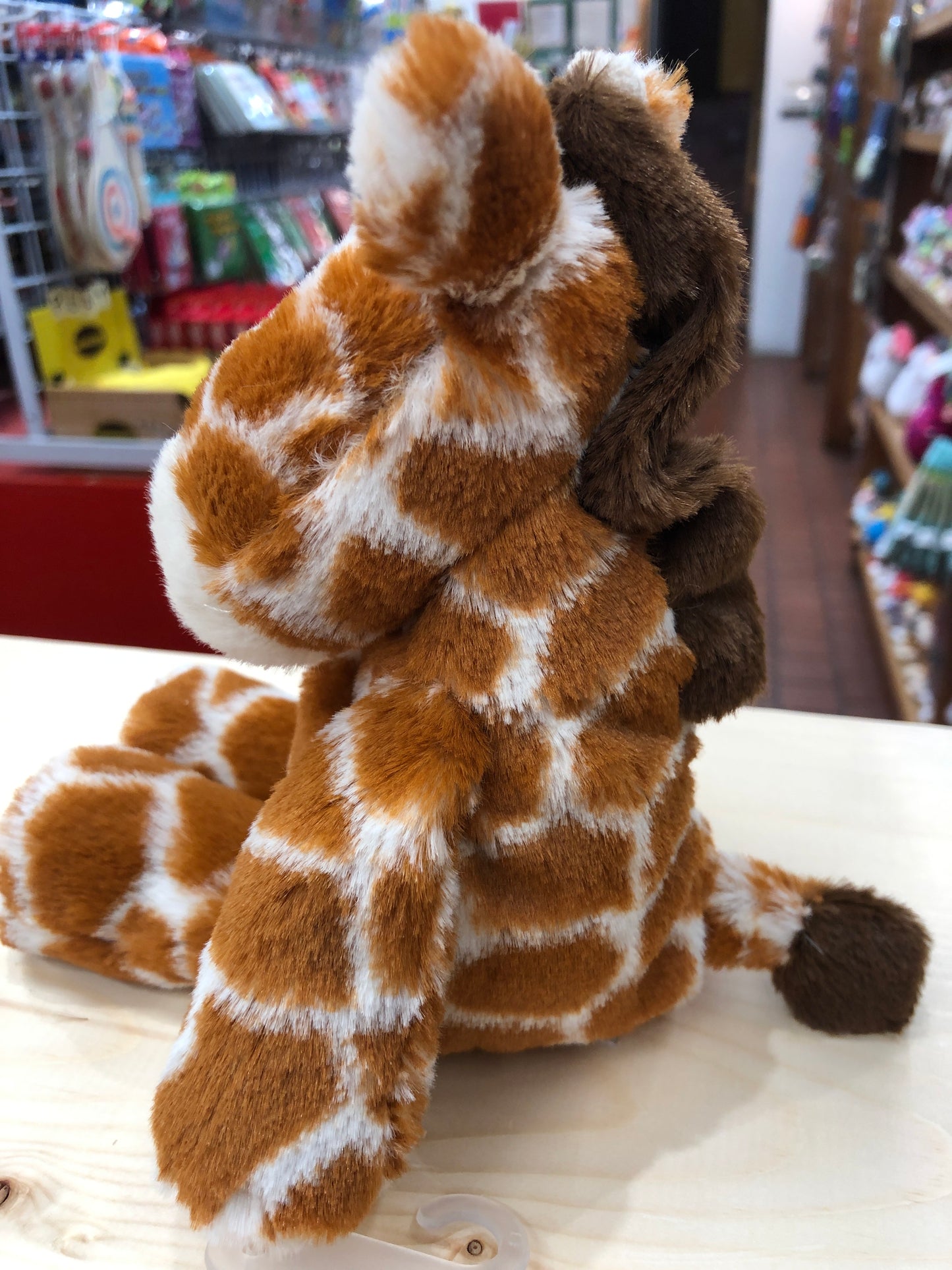 Soft plush giraffe toy from Mary Meyer Marshmallow Zoo, sitting on display in a store with colorful products in background