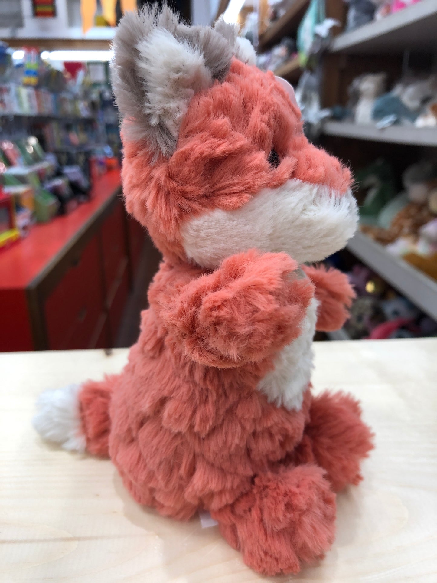 Soft, smiling Mary Meyer Puttling Fox plush toy in warm coral color, sitting on display in a toy store