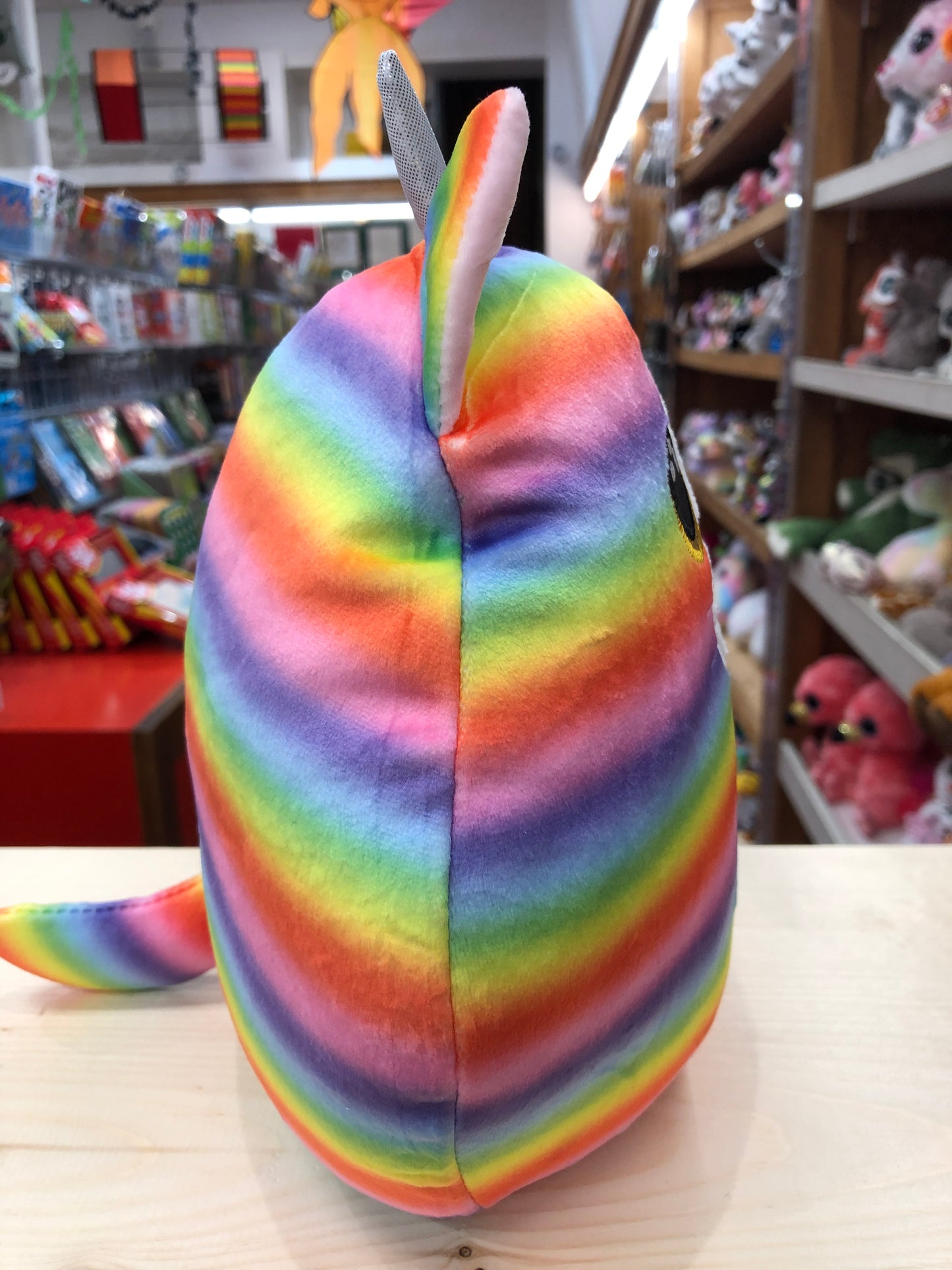 Rainbow striped Ty Squish-a-Boo cat plush toy with ears and tail, displayed in toy store with shelves of stuffed animals