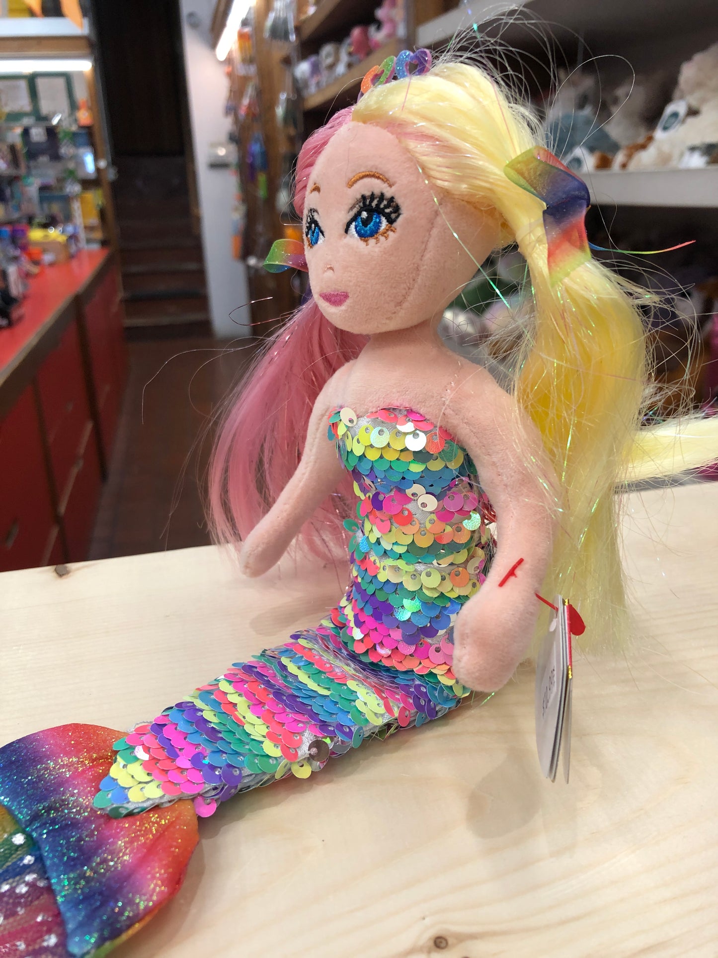 Colorful Anastasia mermaid plush with rainbow sequin tail, two-toned hair, and sparkly crown, sitting on display in toy store