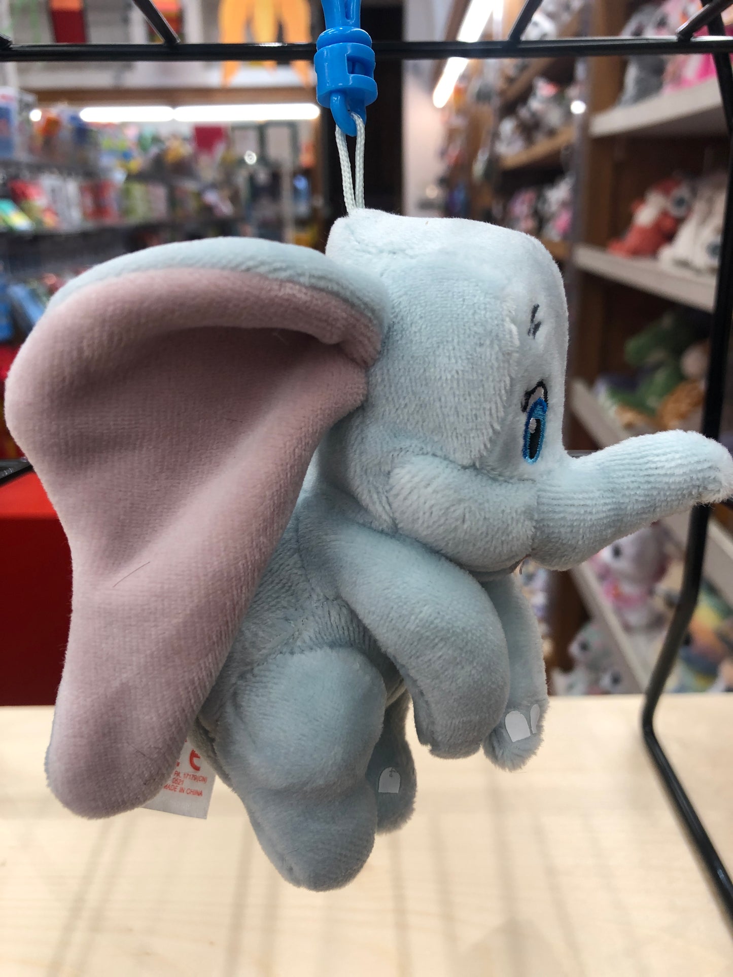 Soft plush Dumbo elephant toy with large ears, hanging from blue clip in toy store display
