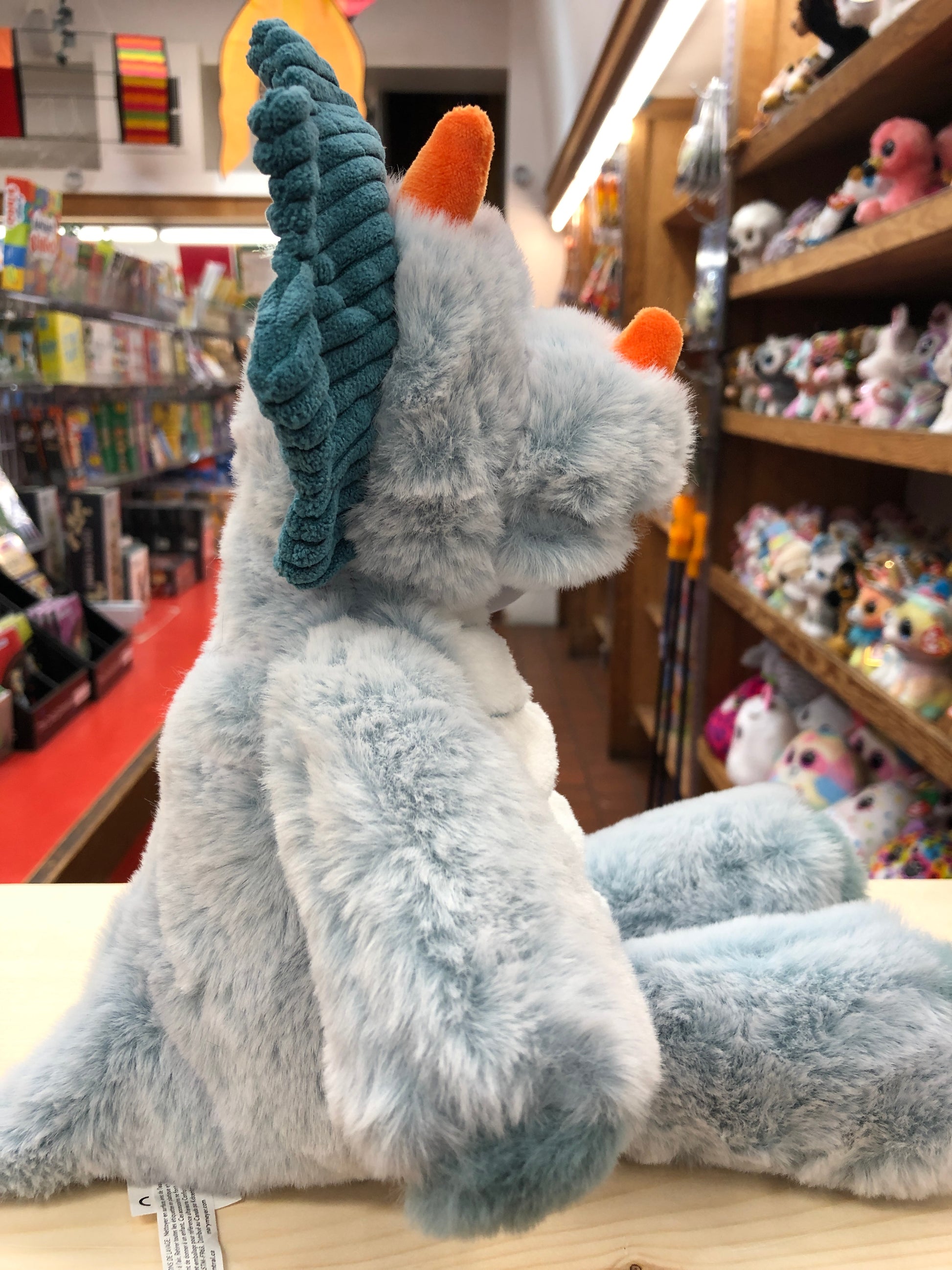 Soft gray Mary Meyer Marshmallow Triceratops plush toy with teal crest in toy store, surrounded by other stuffed animals
