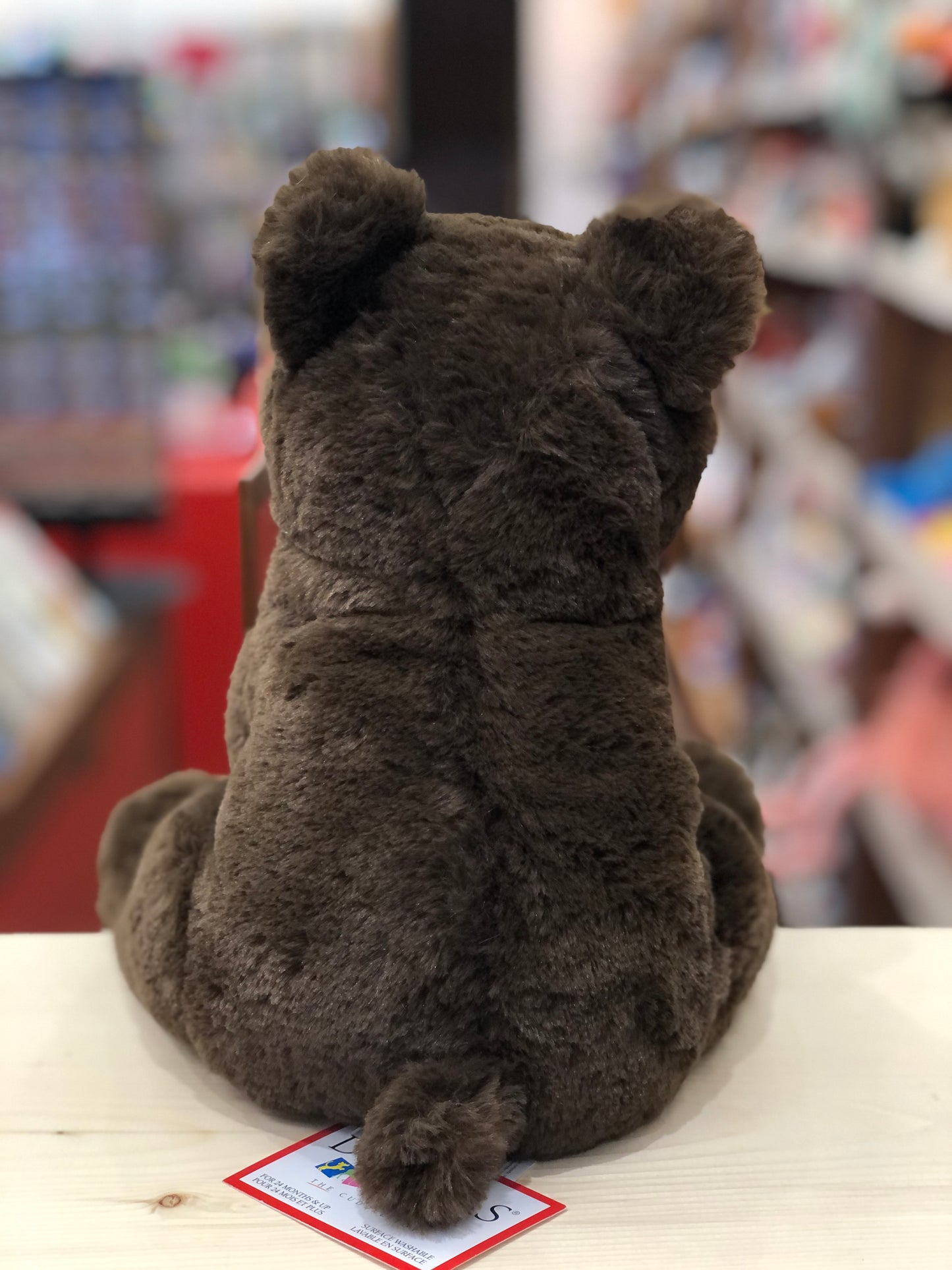 Soft, chocolate brown plush bear cub sitting on display, viewed from behind, showcasing realistic furry texture