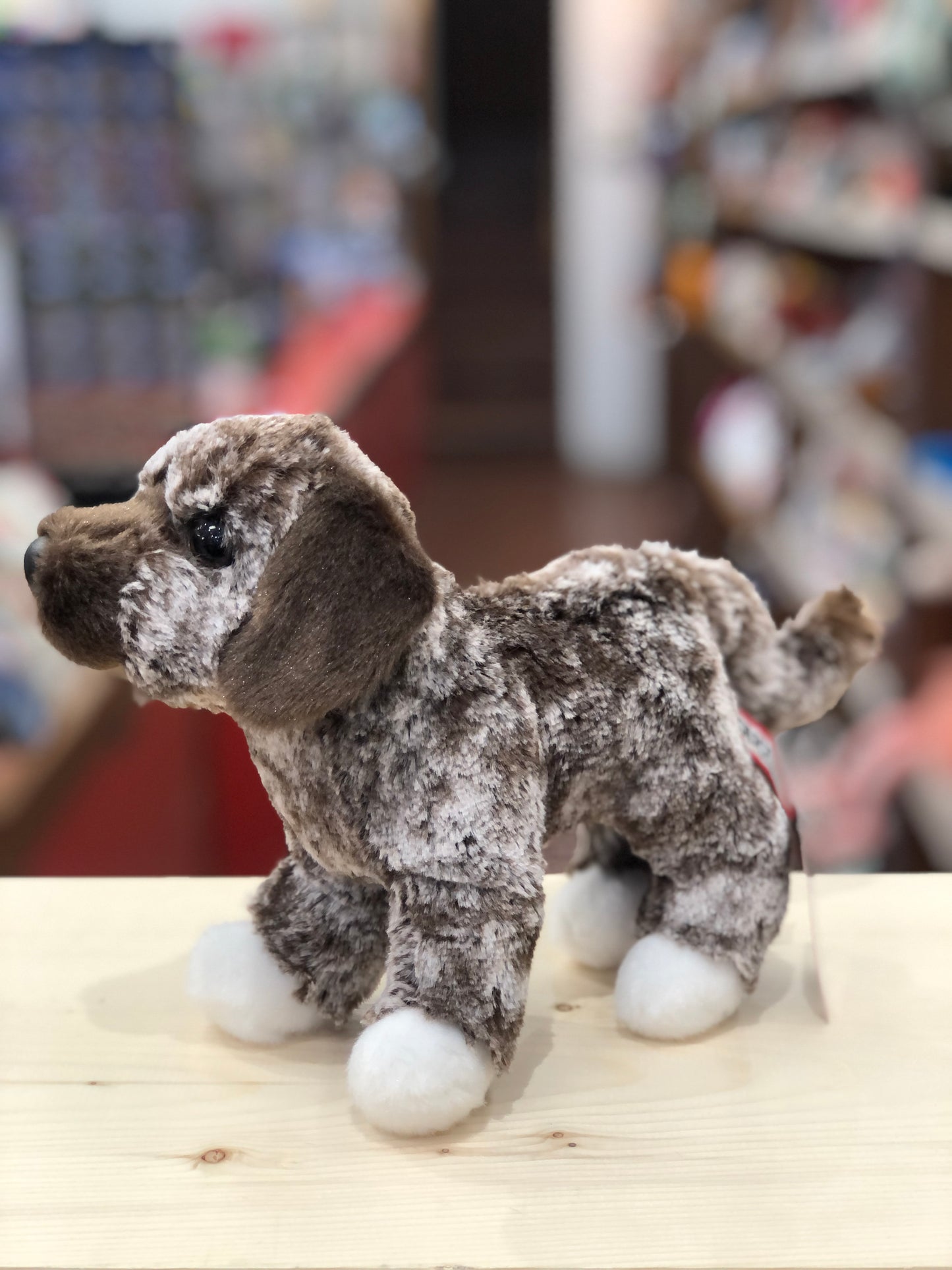 Douglas Spud The Mutt plush toy with brindle coat, white paws, and soulful brown eyes, standing on wooden surface in toy store