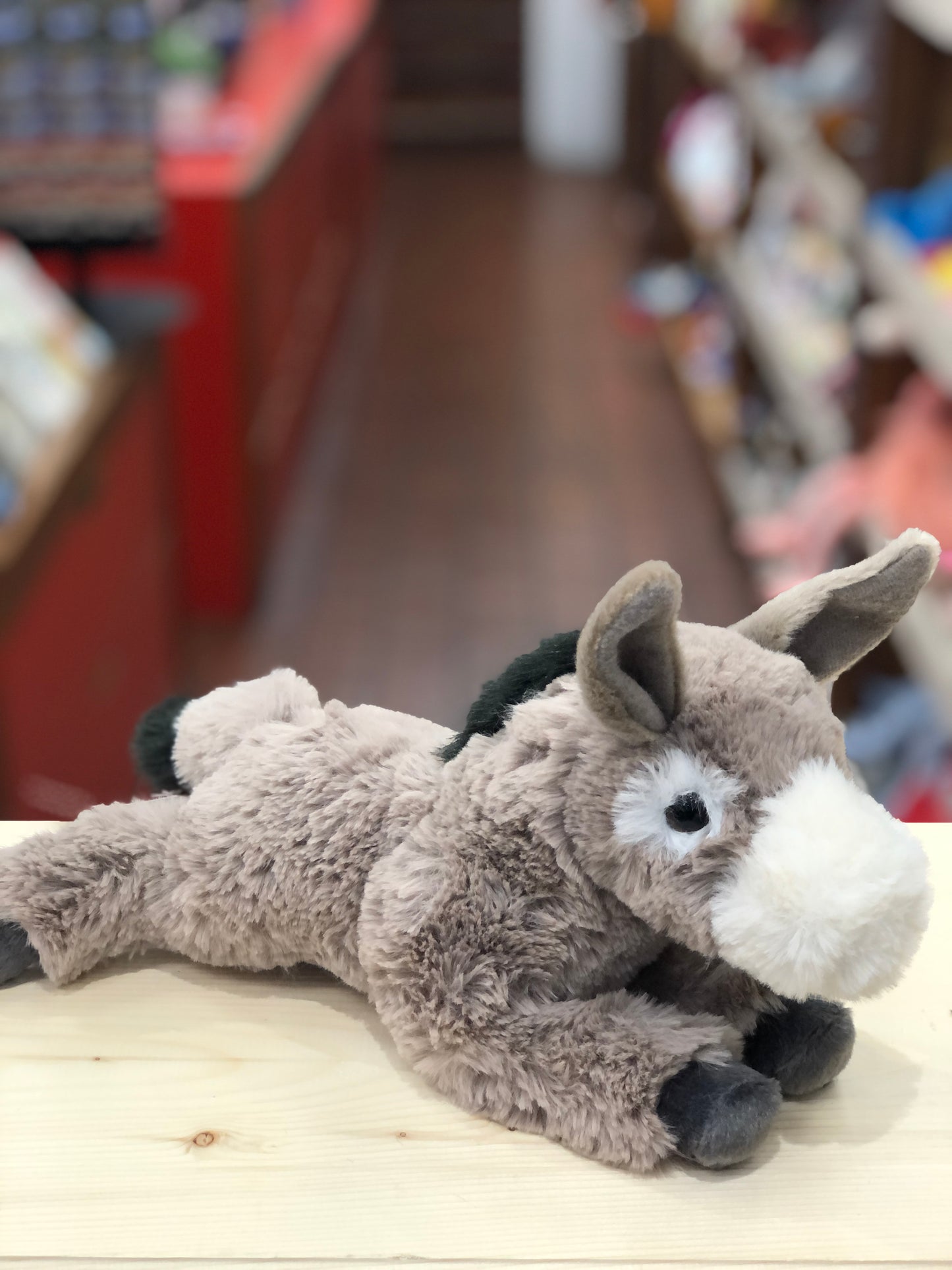 Soft gray floppy donkey plush toy with white face, black hooves and mane, lying on wooden surface in toy store