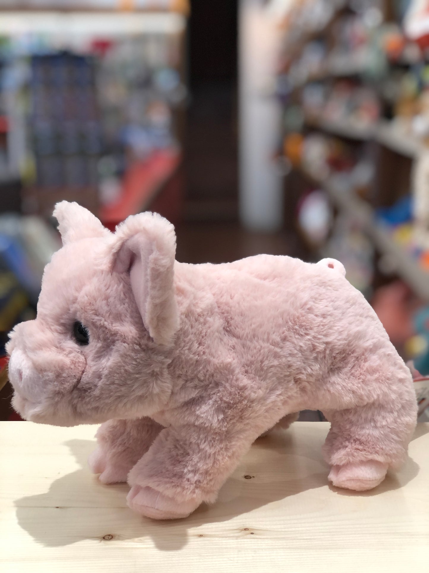 Soft pink pig plush toy with large ears and detailed features, standing on wooden surface in toy store setting