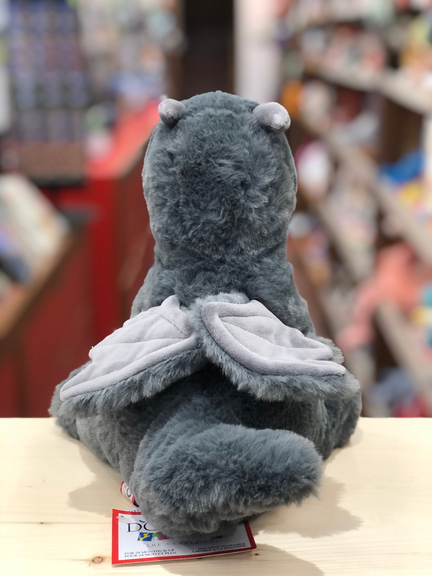 Cuddly gray Sootie dragon plush toy with small wings, sitting on shelf in toy store, back view showing soft textured body