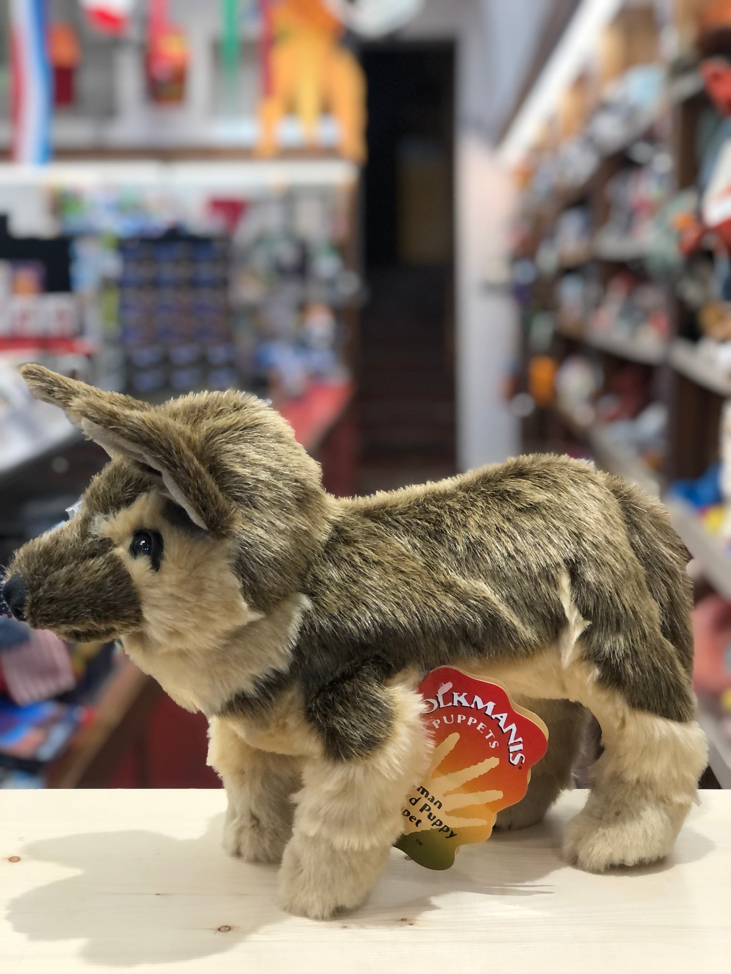 Folkmanis German Shepherd puppy puppet with movable mouth and legs, displayed in toy store with colorful background
