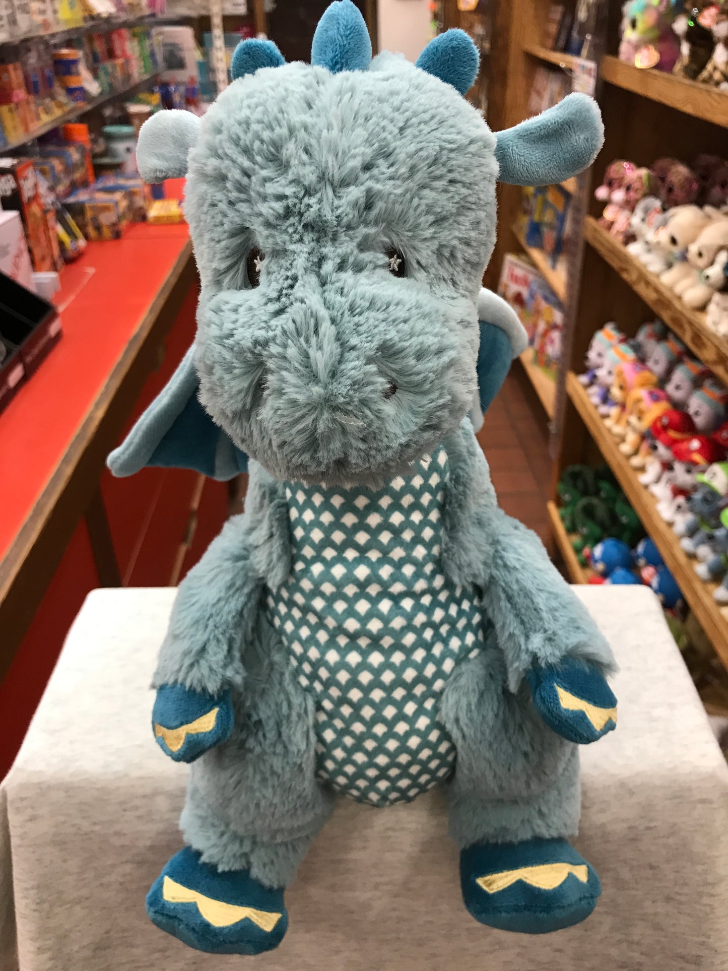 Adorable pastel blue Demitri dragon plush toy with golden claws, patterned belly, and small wings, displayed in a toy store