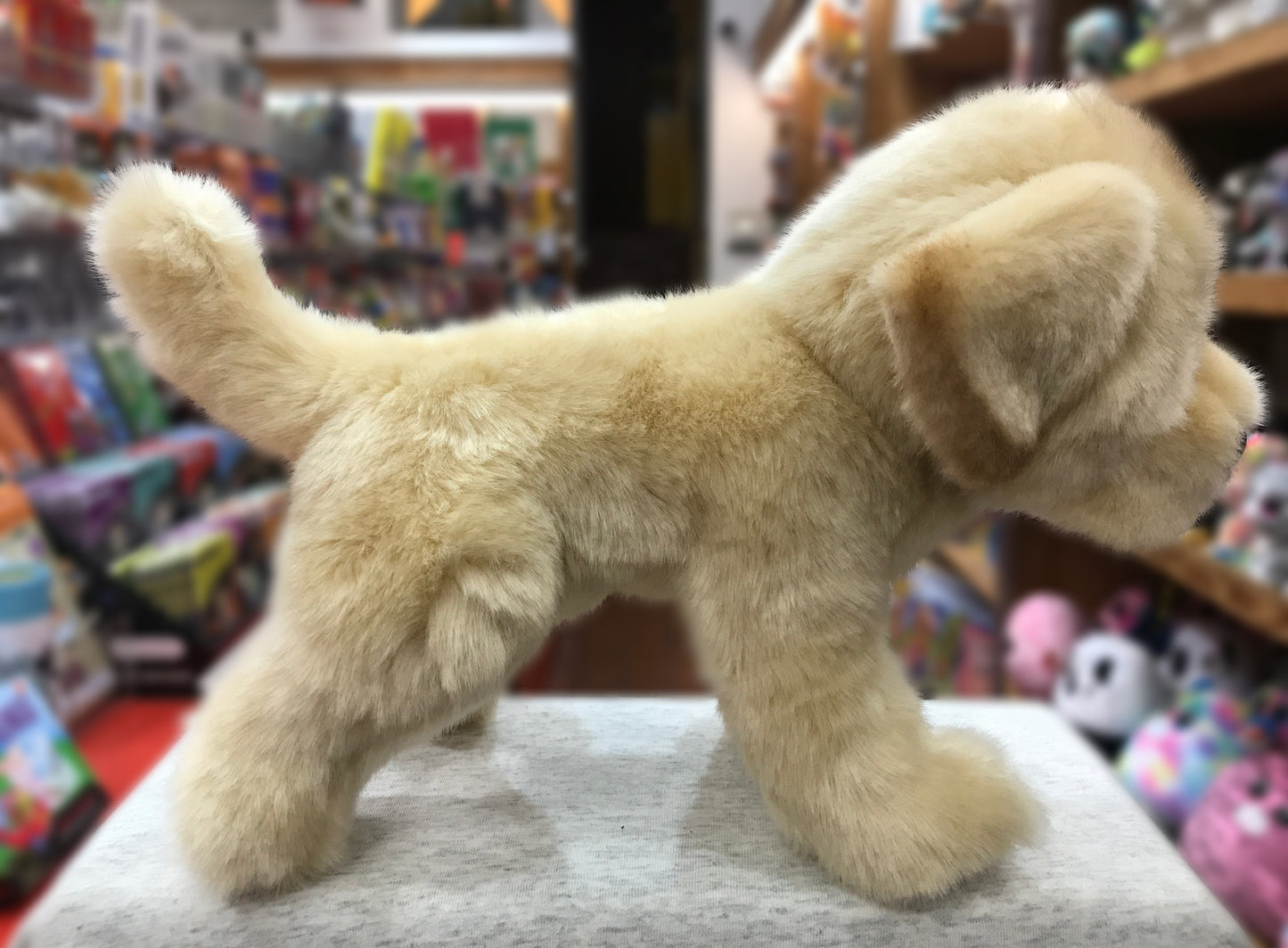 Soft, cuddly yellow Labrador plush toy standing on display in toy store, surrounded by colorful shelves of other stuffed animals