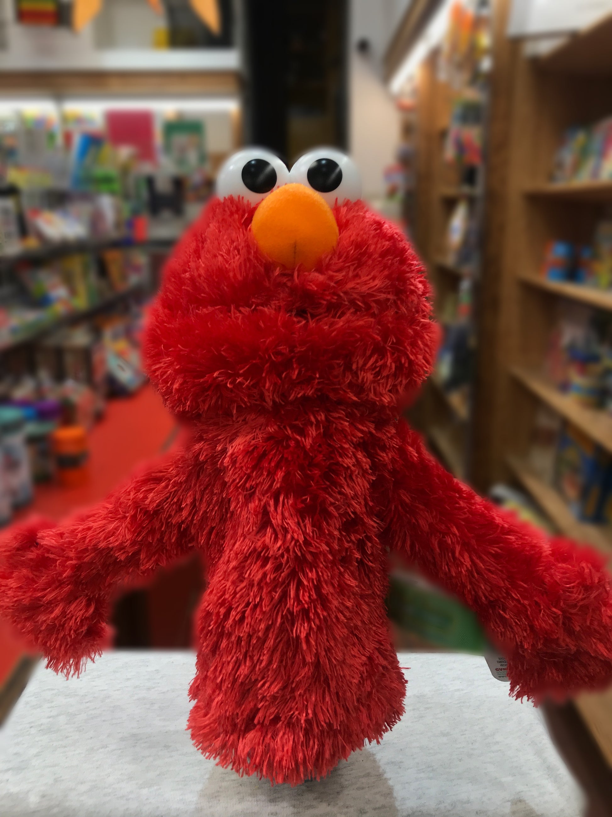 Plush Elmo hand puppet from GUND's Sesame Street collection, displayed in a toy store with bookshelves in background