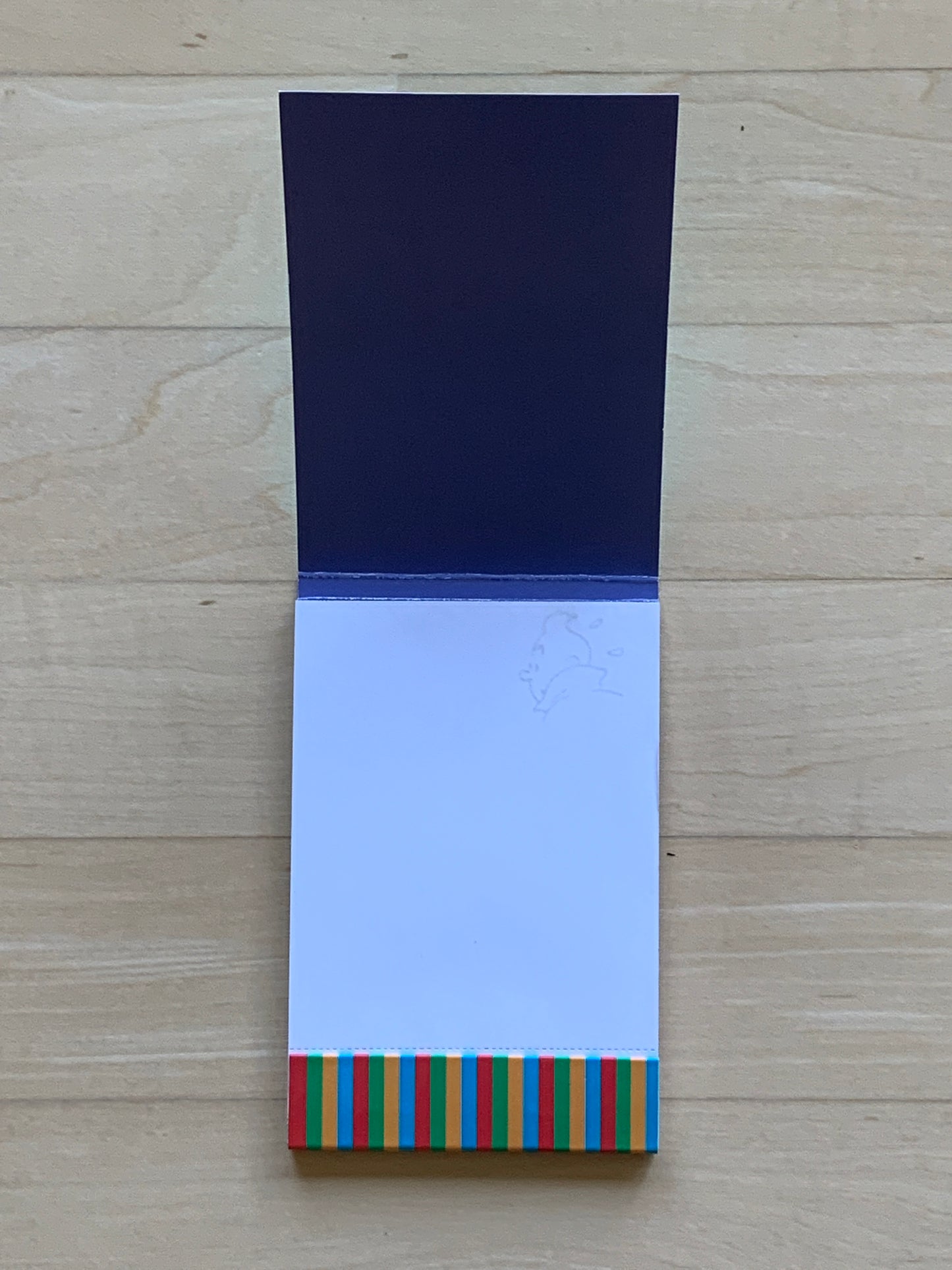 Tintin notepad with navy cover, watermarked pages, and colorful striped bottom edge, open to reveal blank page