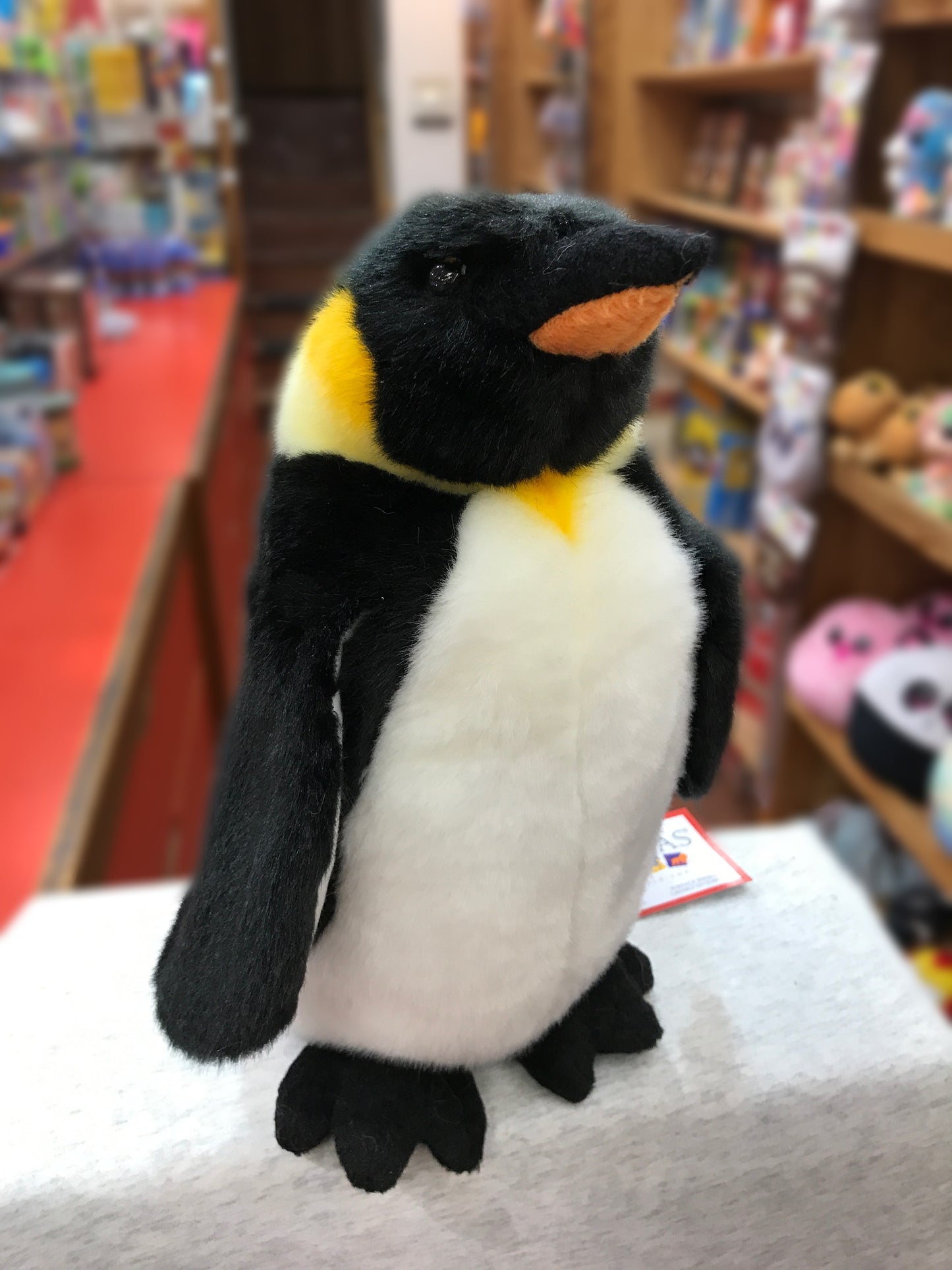 Soft plush Emperor Penguin toy with realistic black and white coloring and orange-yellow neck details, displayed in a toy store