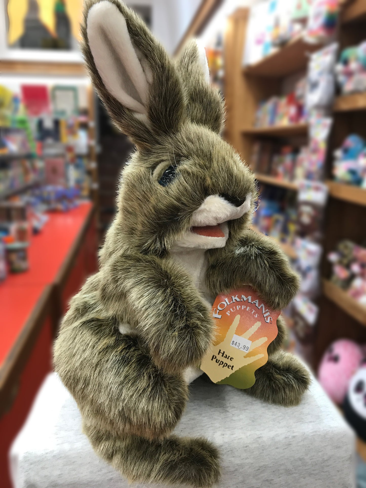 Folkmanis Hare Puppet with movable mouth and forelegs, standing tall in a toy store, displaying its realistic fur and expressive face