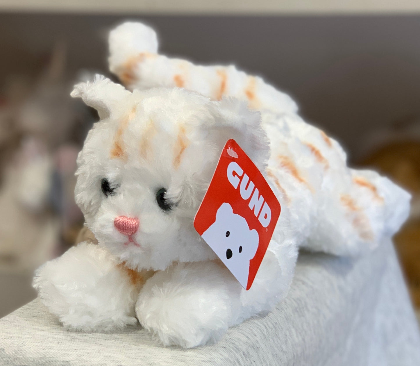 Gund Bootsie white cat plush toy with orange stripes, pink nose, and red brand tag, sitting on soft surface