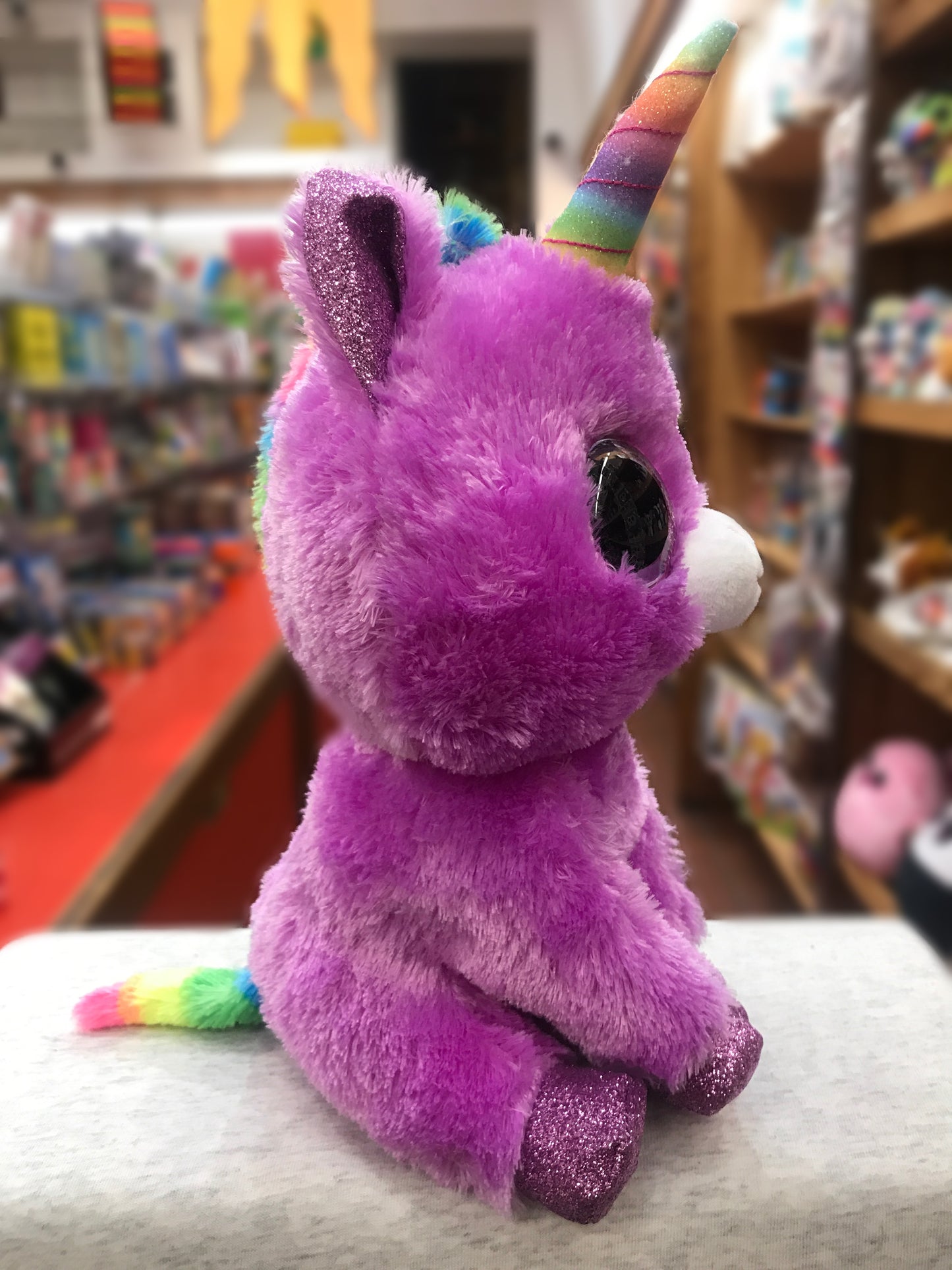 Ty Beanie Boo Rosette unicorn plush, purple with rainbow horn and tail, sparkly accents, sitting in bookstore display