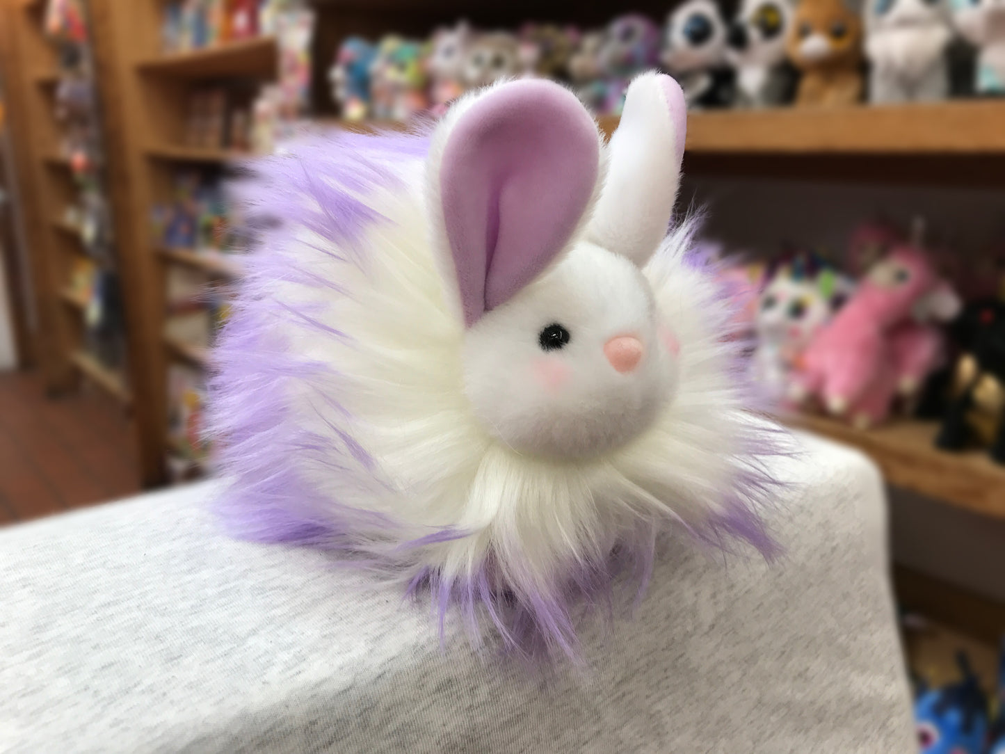 Fluffy white and purple bunny plush toy with long fur, pink nose, and large ears, displayed in a toy store setting