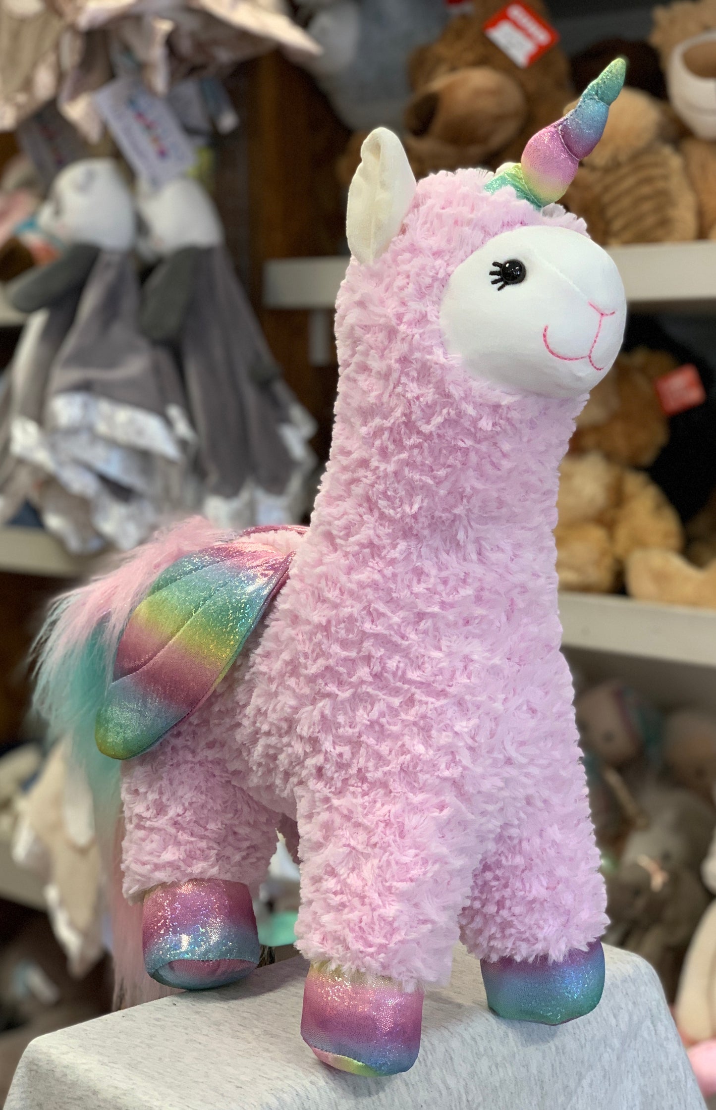 Gund Llamacorn plush toy with pink fur, rainbow horn and hooves, sparkly wings, and smiling face, surrounded by other stuffed animals