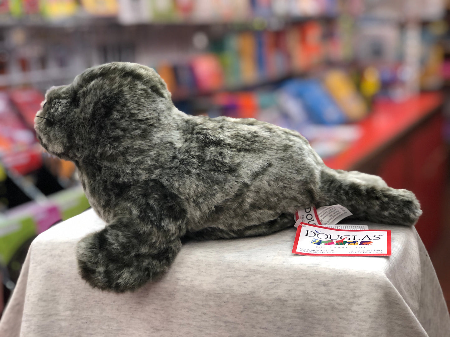Realistic Douglas Speckles monk seal plush toy with silvery fur, displayed in colorful toy store setting