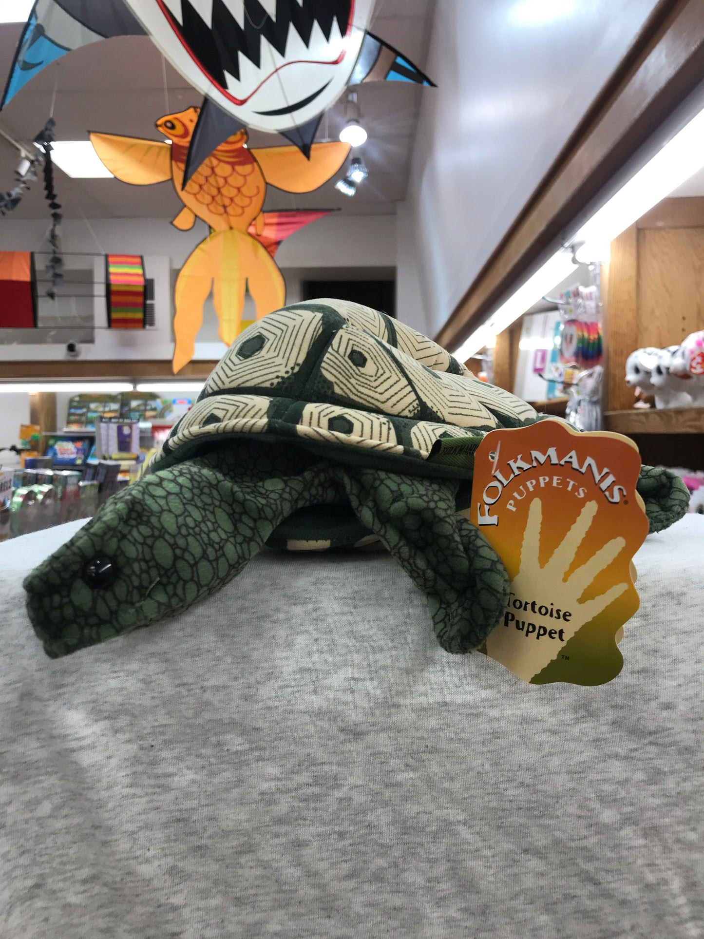 Folkmanis Tortoise Puppet on display in toy store, with patterned shell and Folkmanis tag visible. Colorful hanging decorations in background.