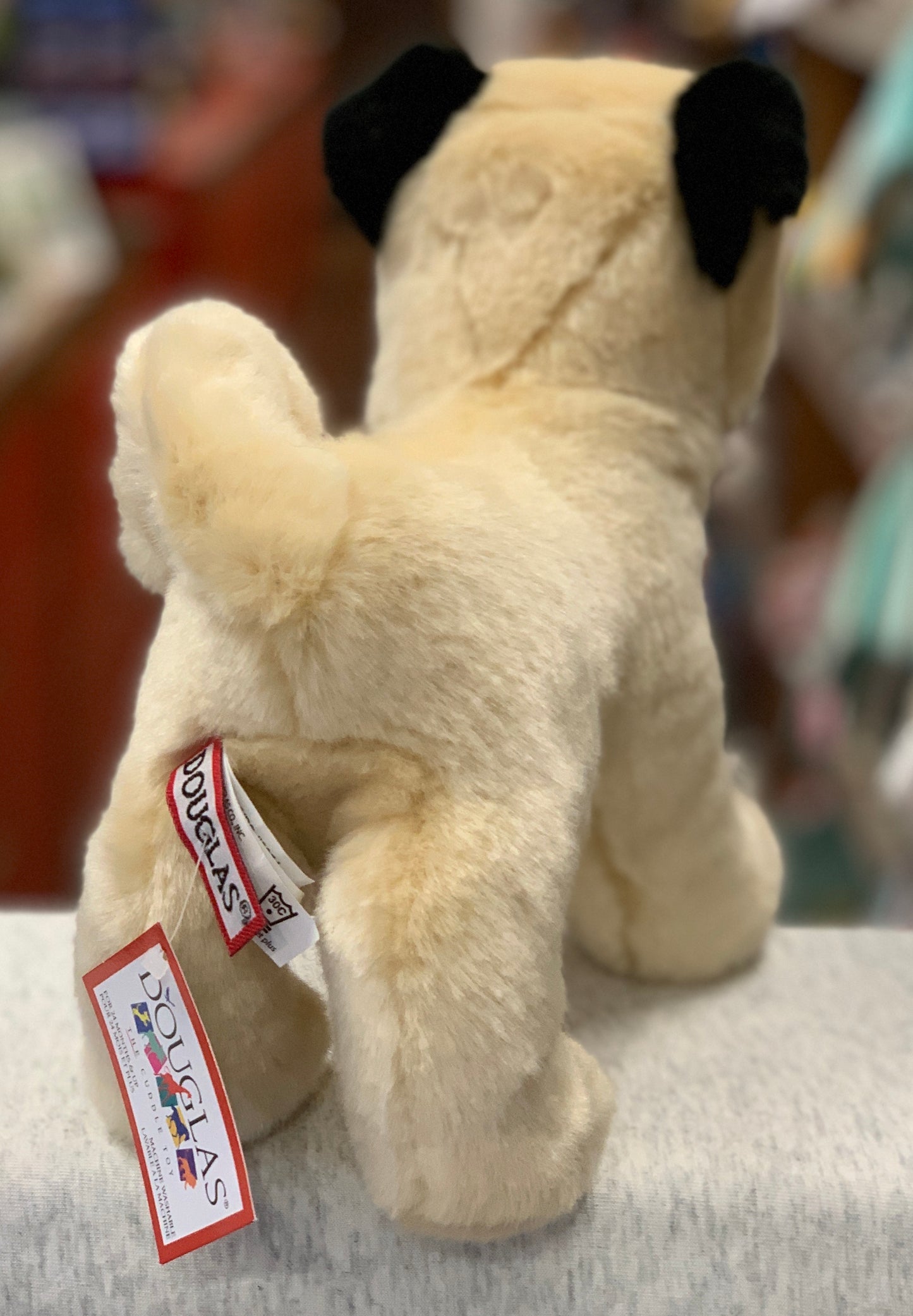 Cute plush pug dog toy, tan with black ears, standing upright with Douglas brand tag, soft and cuddly 10-inch stuffed animal