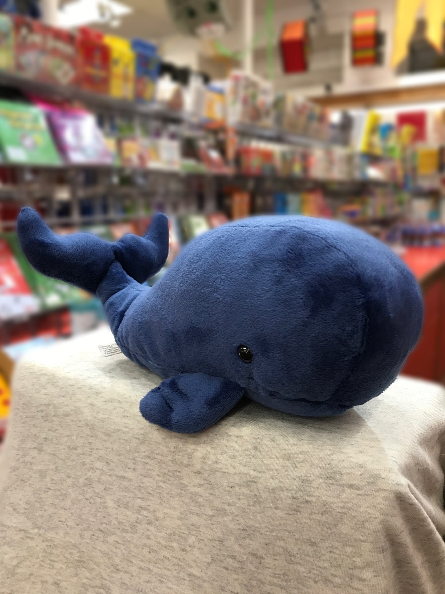 Soft navy blue whale plush toy with friendly eyes and tail, displayed in colorful toy store setting