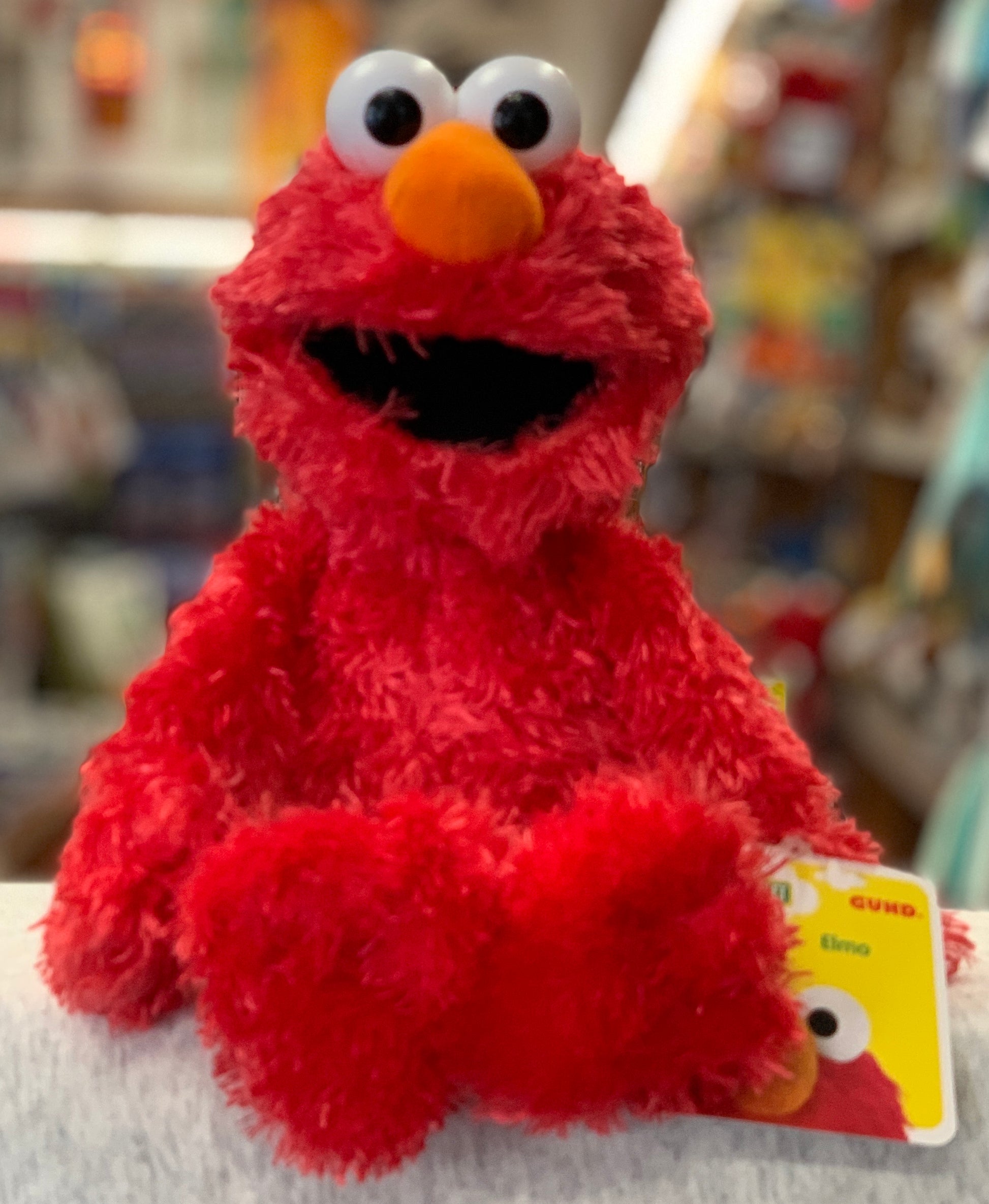 Gund Sesame Street Elmo plush toy, 13 inches tall, with bright red fur, large eyes, and orange nose, displayed in store