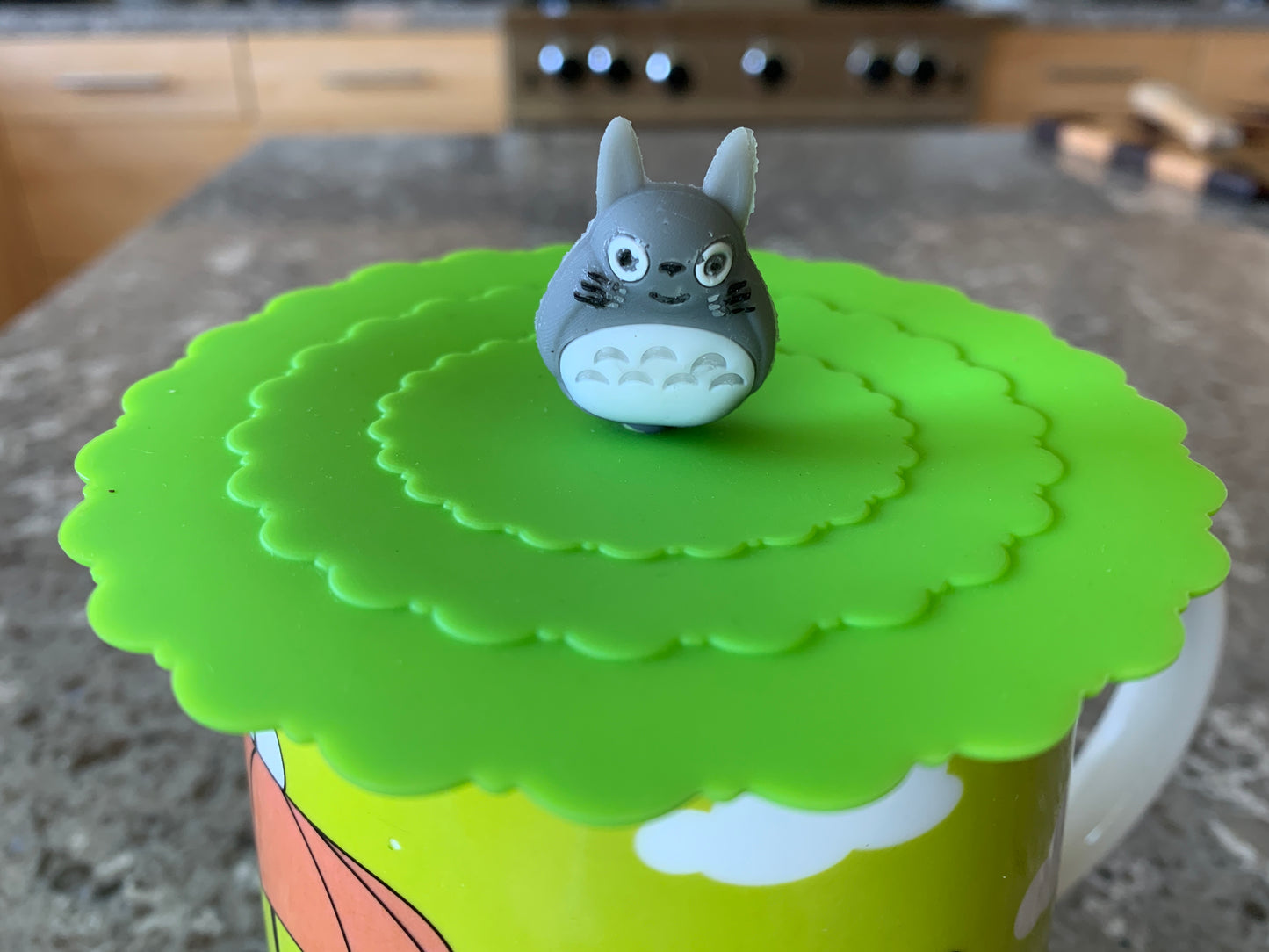 Smiling Totoro figure on bright green silicon cup cover, placed on colorful mug on kitchen counter