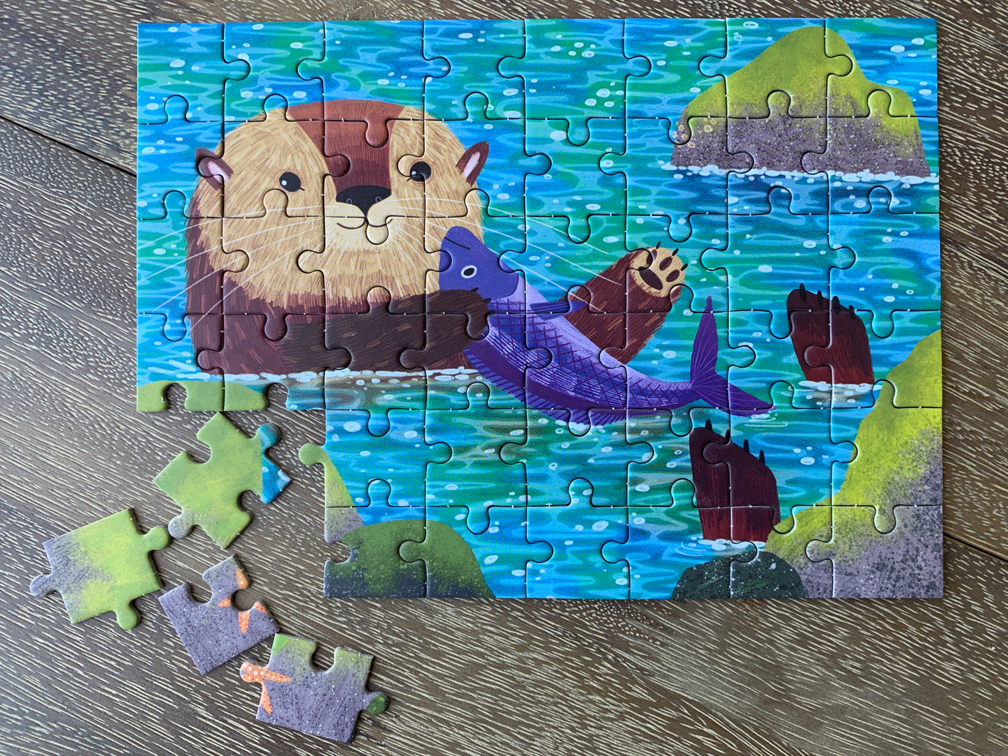 Colorful otter puzzle with sea scene, showing cute otter holding purple fish. Some pieces removed, revealing gift set contents.