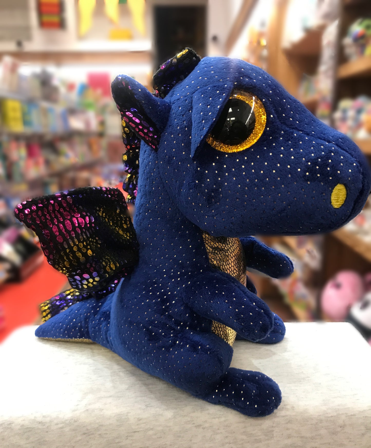 Ty Beanie Boo Saffire blue dragon plush with gold speckles and colorful wings, large glittery eyes, in a bookstore setting