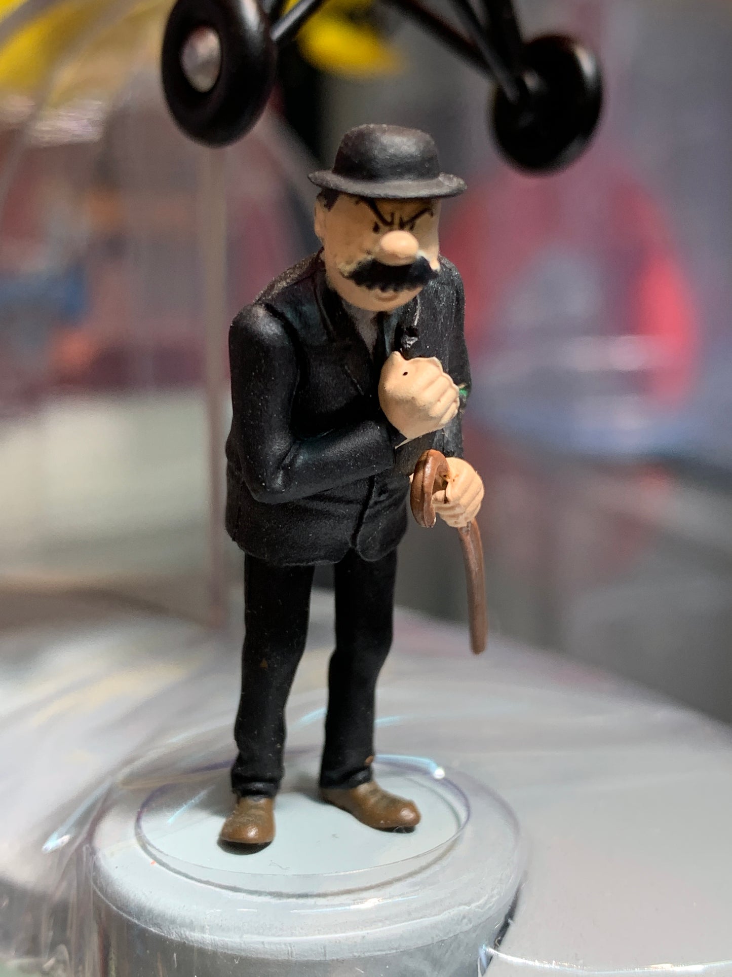 Miniature figurine of a mustachioed man in black suit and hat holding a cane, displayed on a white stand