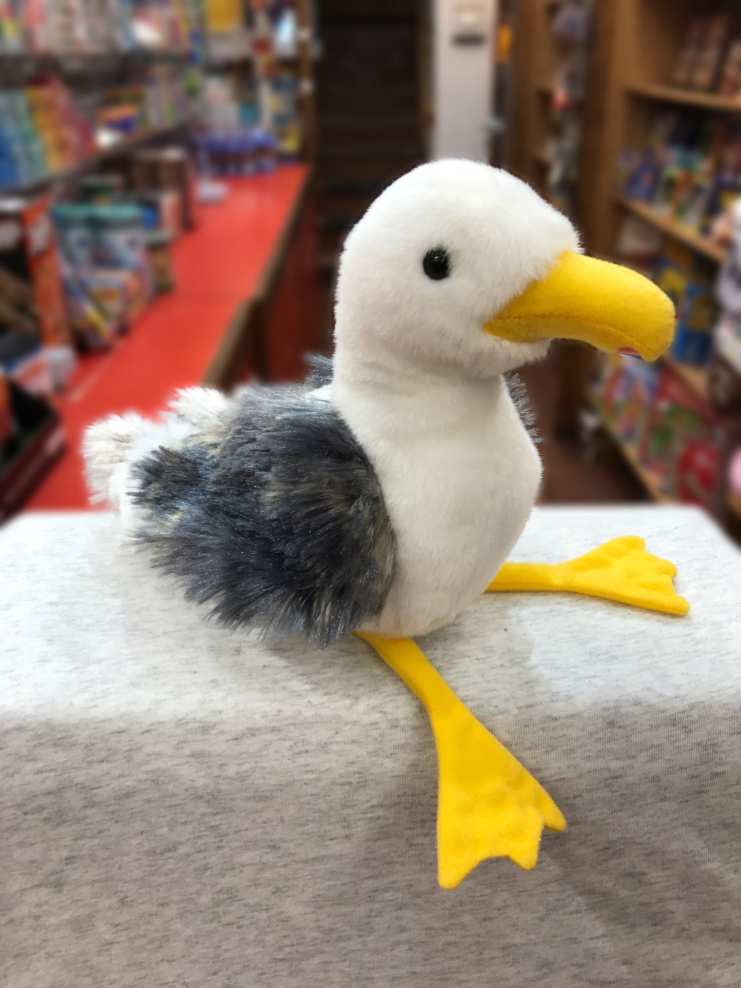 Soft plush seagull toy with white body, gray wings, yellow beak and feet, displayed in a toy store setting