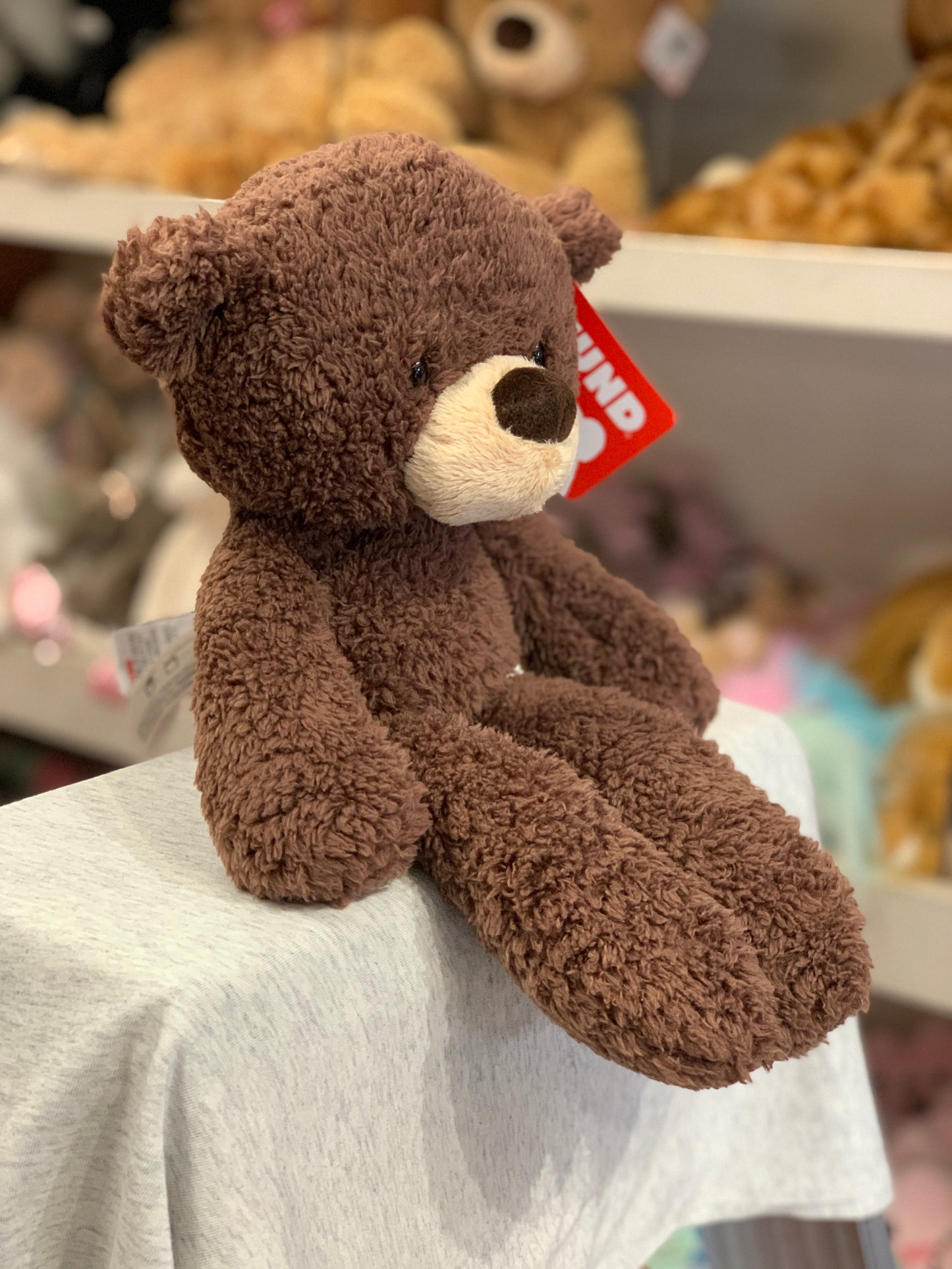 Gund Fuzzy Bear plush toy in chocolate brown, sitting on display shelf with other stuffed animals visible in background