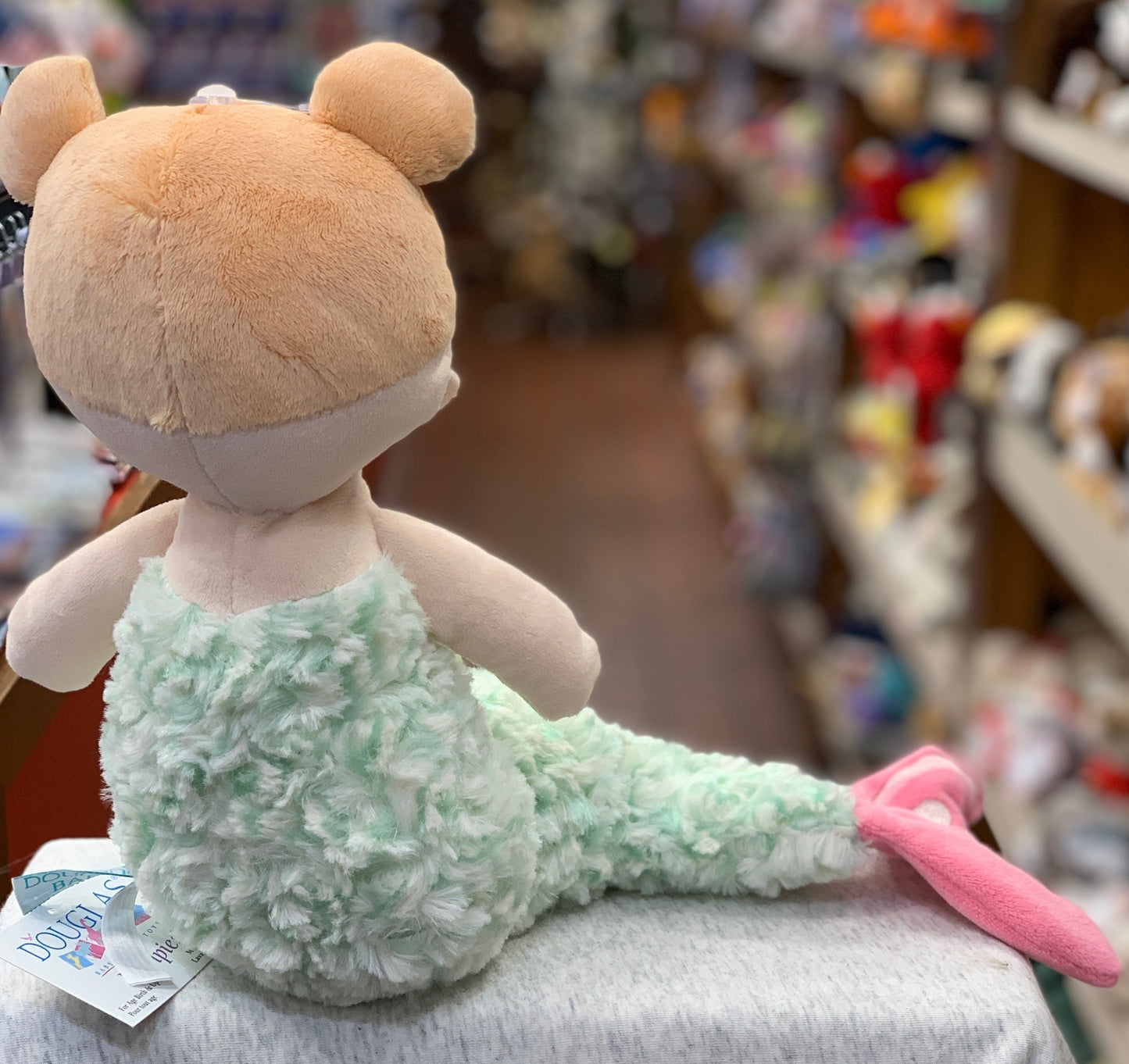 Soft plush mermaid toy with mint green tail, pink fin, and round beige head, sitting on display in toy store