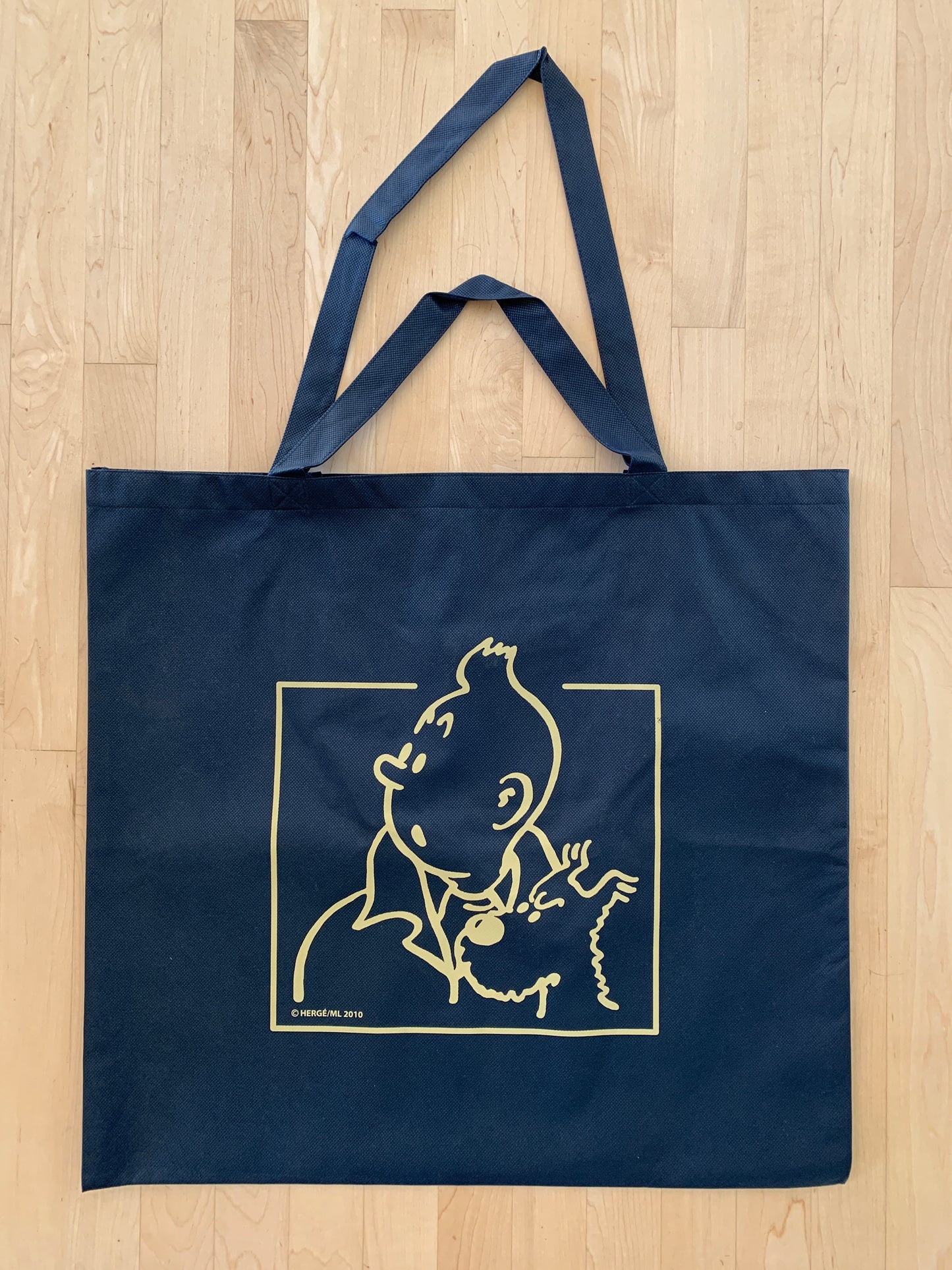 Tintin recycled plastic blue tote bag with gold logo and dual handle sets, short and long, on wooden background