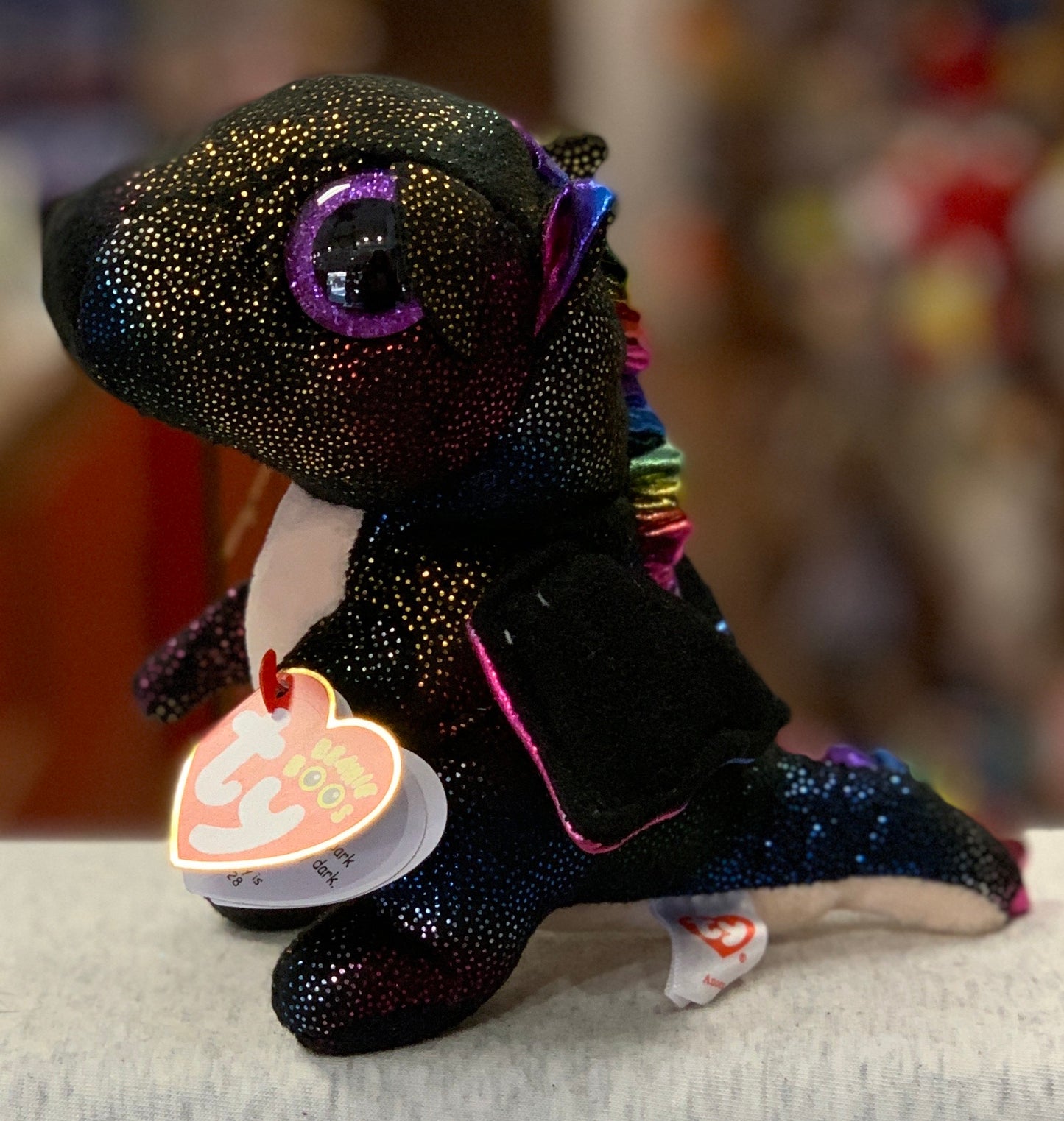 Sparkly black Ty Beanie Boo Anora dragon plush with colorful details and large purple eyes, displaying heart-shaped tag