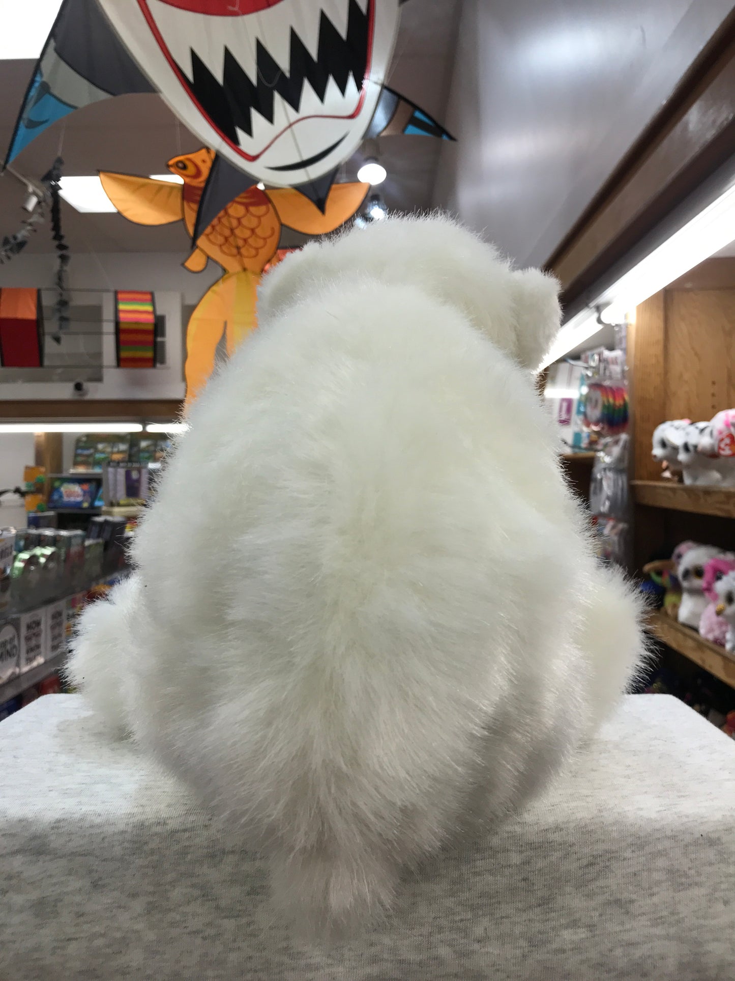 Fluffy white Folkmanis polar bear cub puppet in toy store, surrounded by colorful hanging decorations and shelves of toys