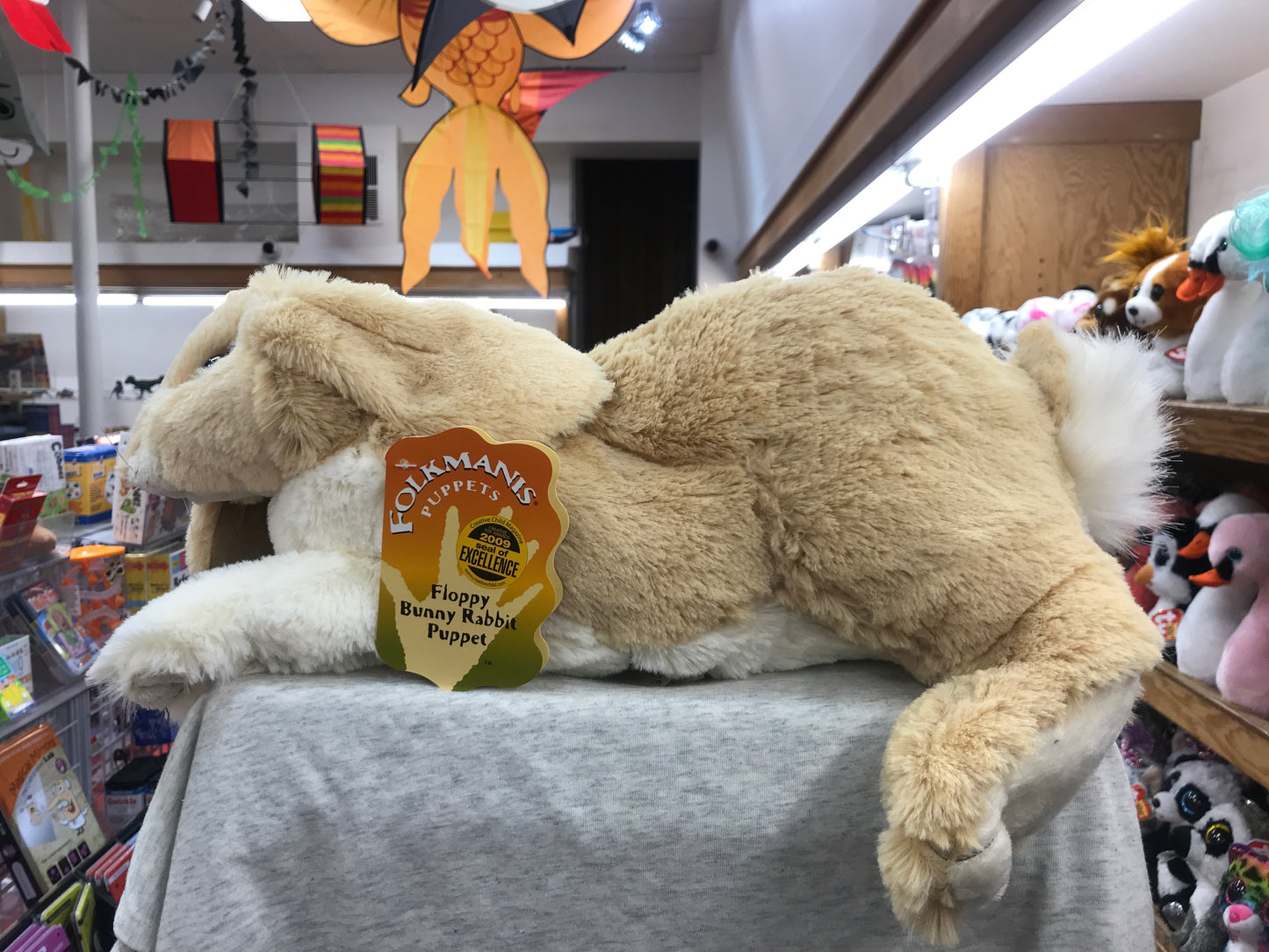Folkmanis Floppy Bunny Rabbit puppet, 17 inches long, soft cream-colored plush toy displayed in toy store with colorful decorations