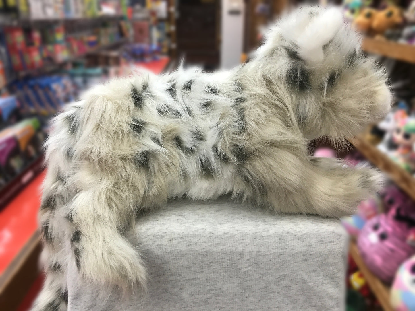 Folkmanis Snow Leopard Cub puppet with spotted plush fur and long tail, displayed in toy store surrounded by colorful toys
