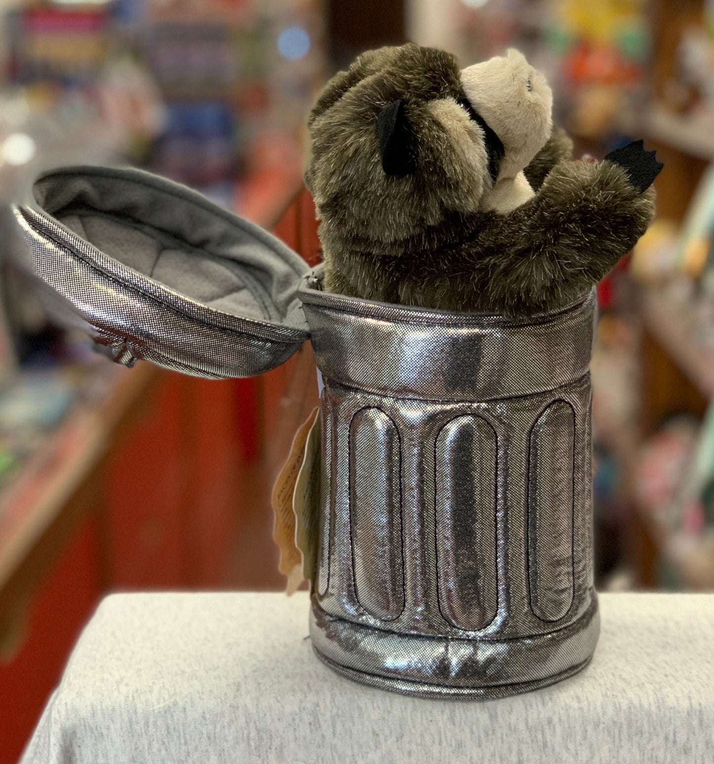 Folkmanis raccoon puppet peeking out of realistic silver garbage can, lid open, in toy store setting