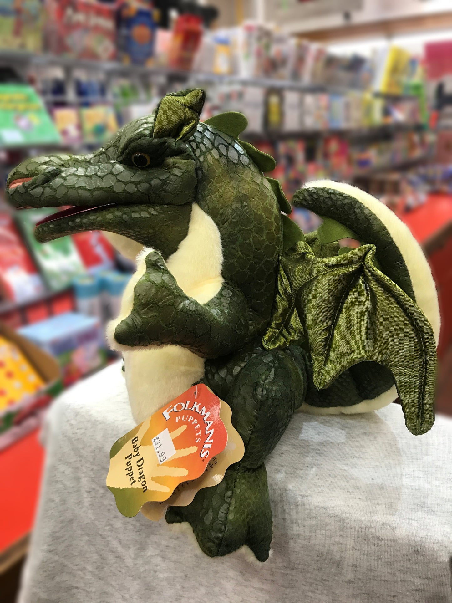 Green Folkmanis Baby Dragon puppet with scaly texture and satin wings, sitting on display in toy store