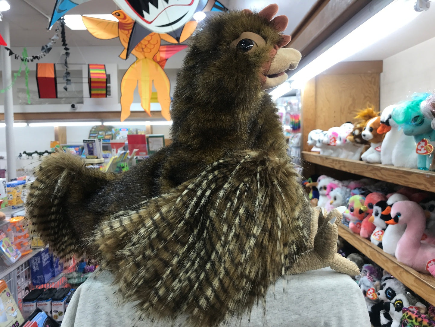Large plush hen puppet in toy store, with feathered brown body and movable beak, surrounded by other stuffed animals